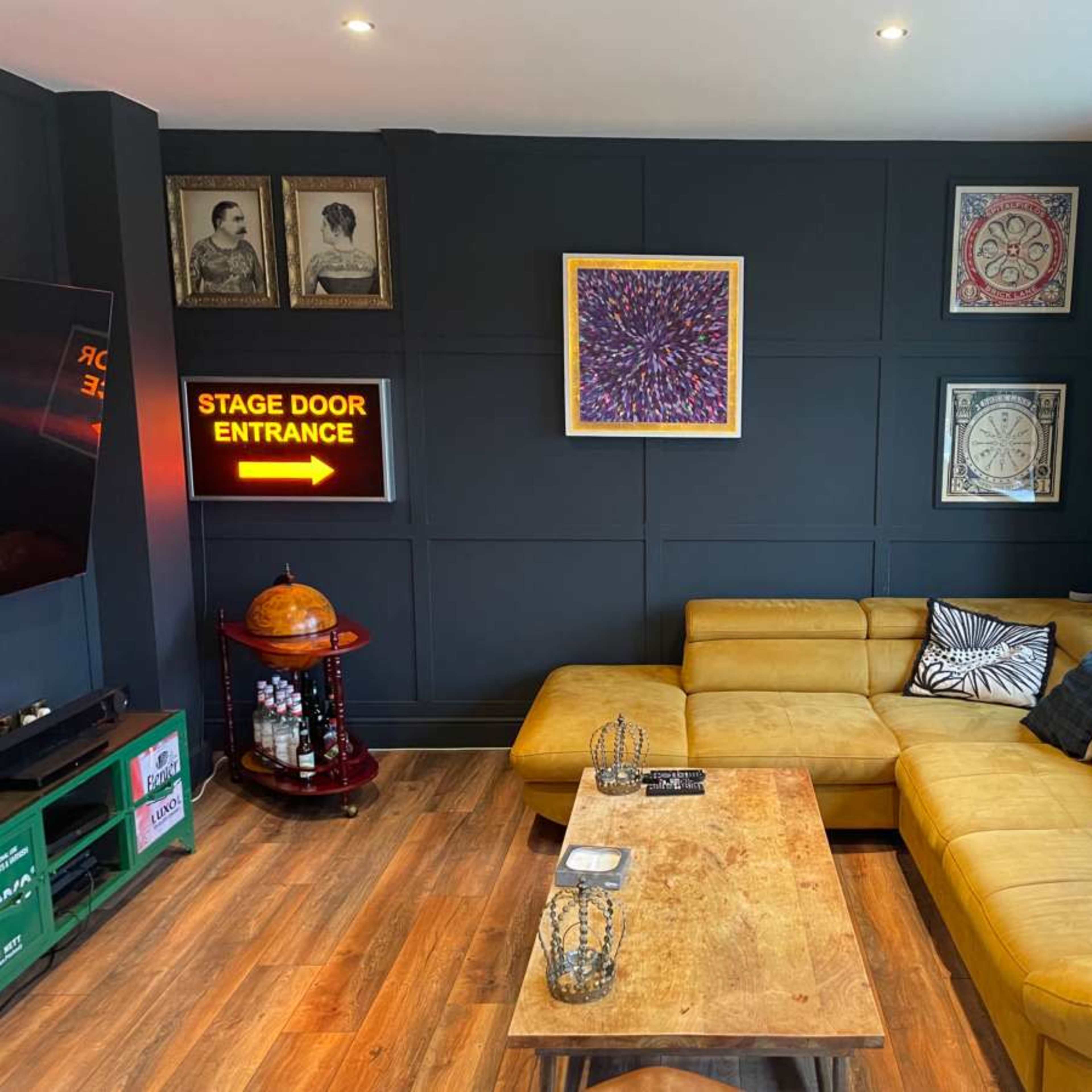 A cozy living room features a yellow sectional sofa, a wooden coffee table, and a wall decorated with framed artwork and a neon sign that reads "STAGE DOOR ENTRANCE."