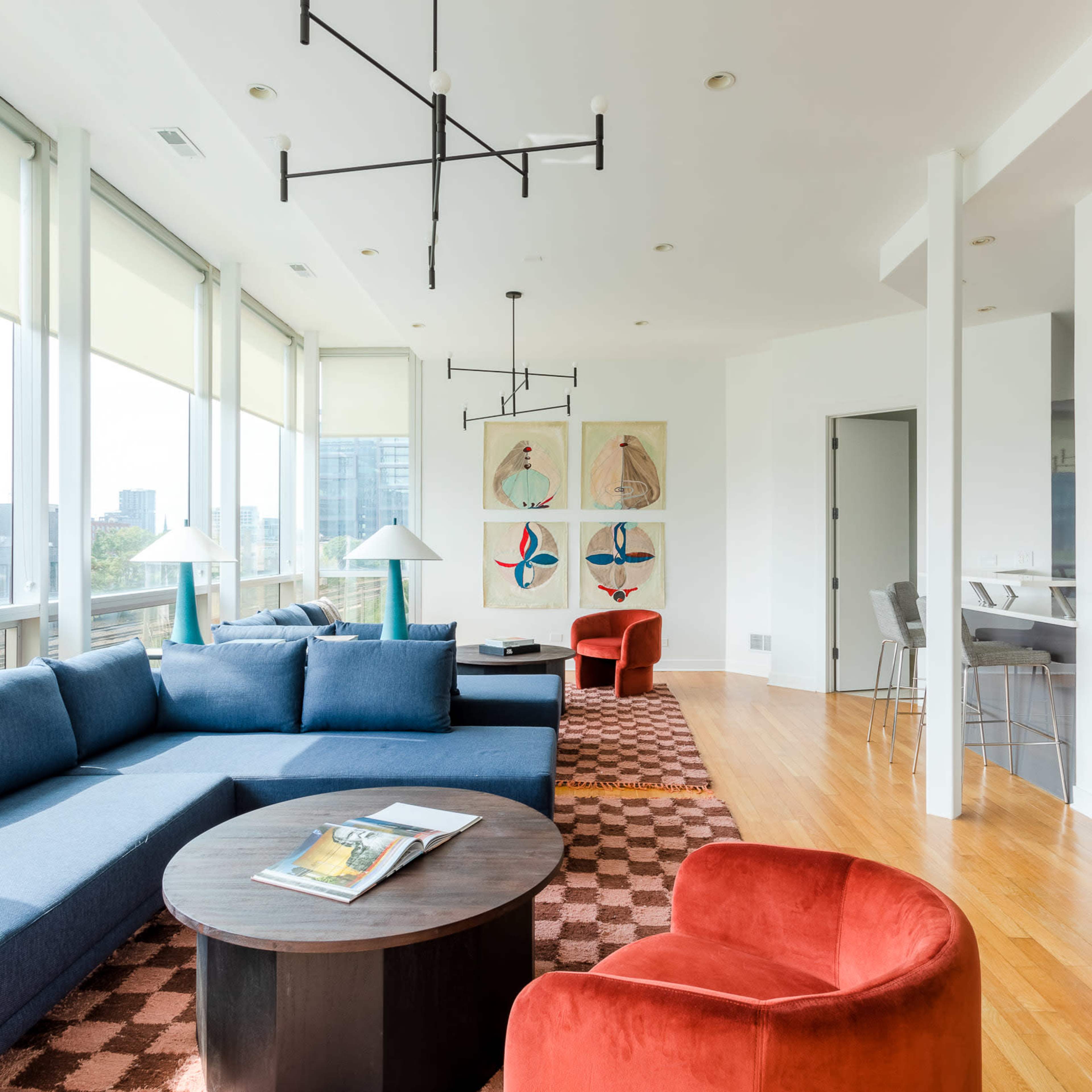 A modern living room with large windows, a blue sectional sofa, a round coffee table, and two red armchairs, featuring abstract art on the walls.