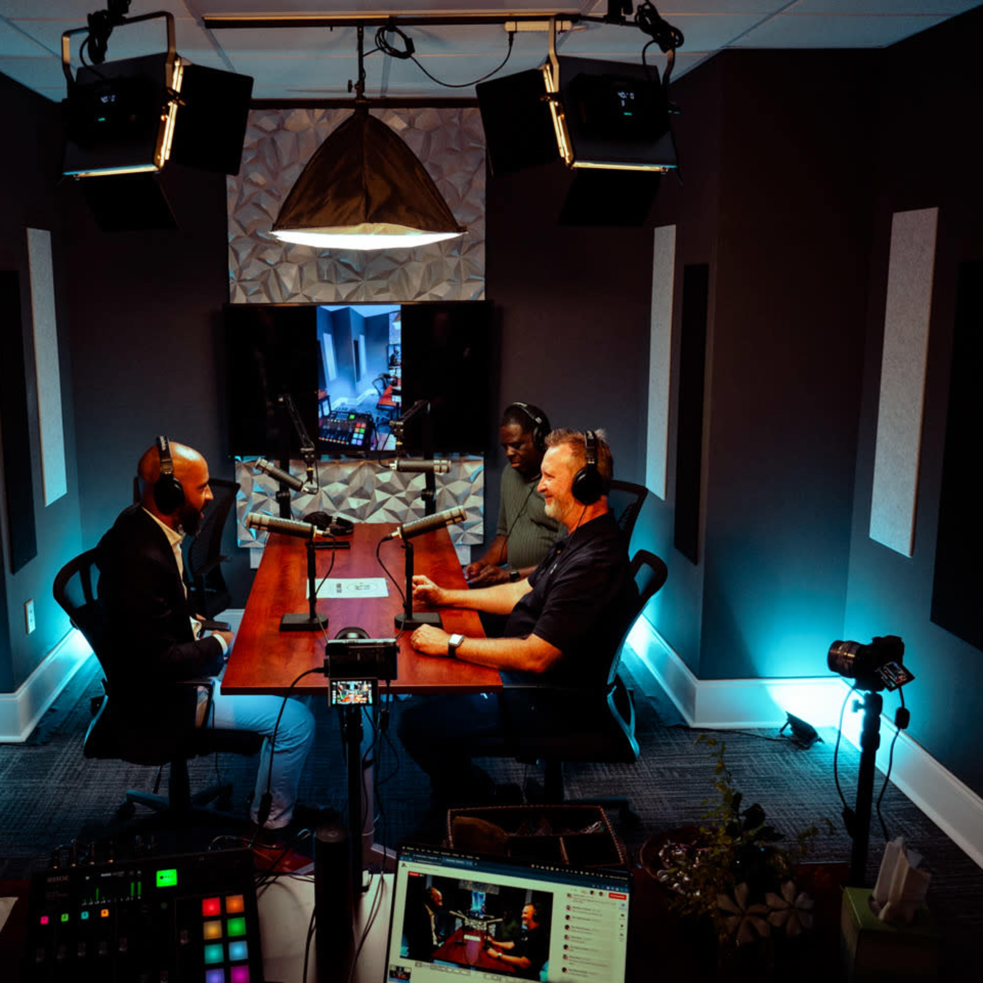 Three people sit around a wooden table in a modern podcast studio, equipped with microphones and camera equipment, while a screen in the background displays a digital interface.