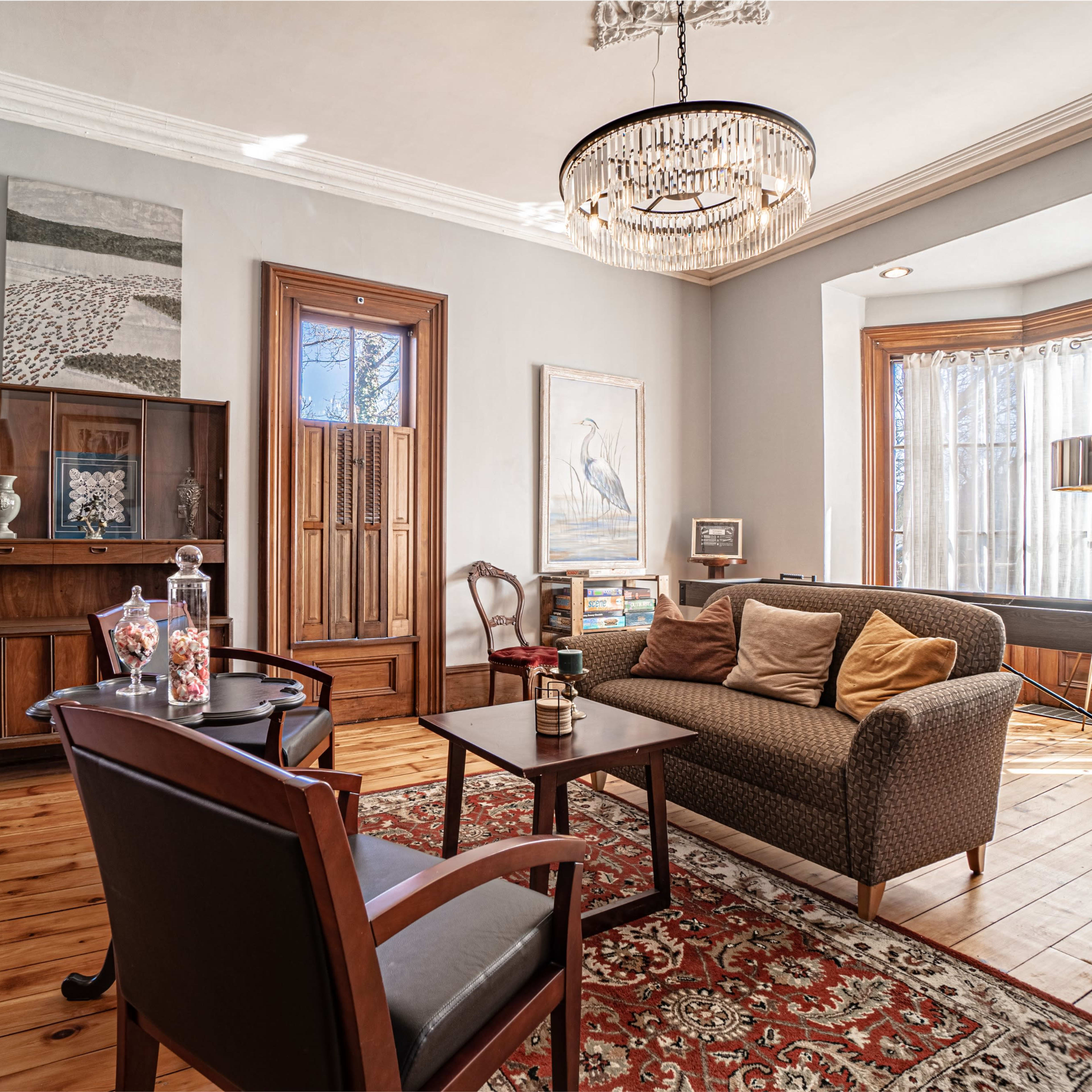 The living room features a sofa with cushions, a wooden cabinet, a chandelier, and large windows with sheer curtains.