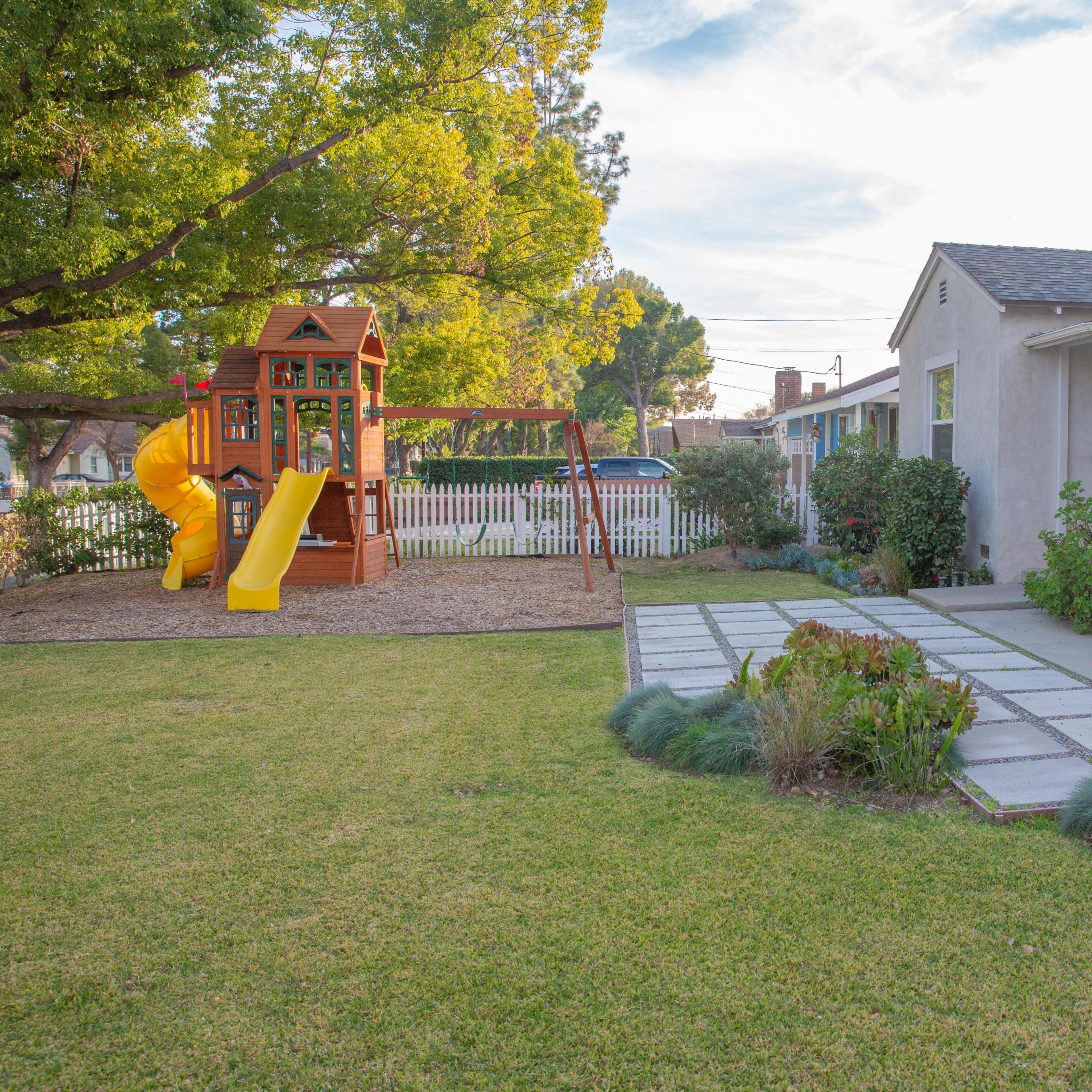 A backyard with a playset featuring a yellow slide and swing set, surrounded by a grassy area and a stone pathway leading to a house.