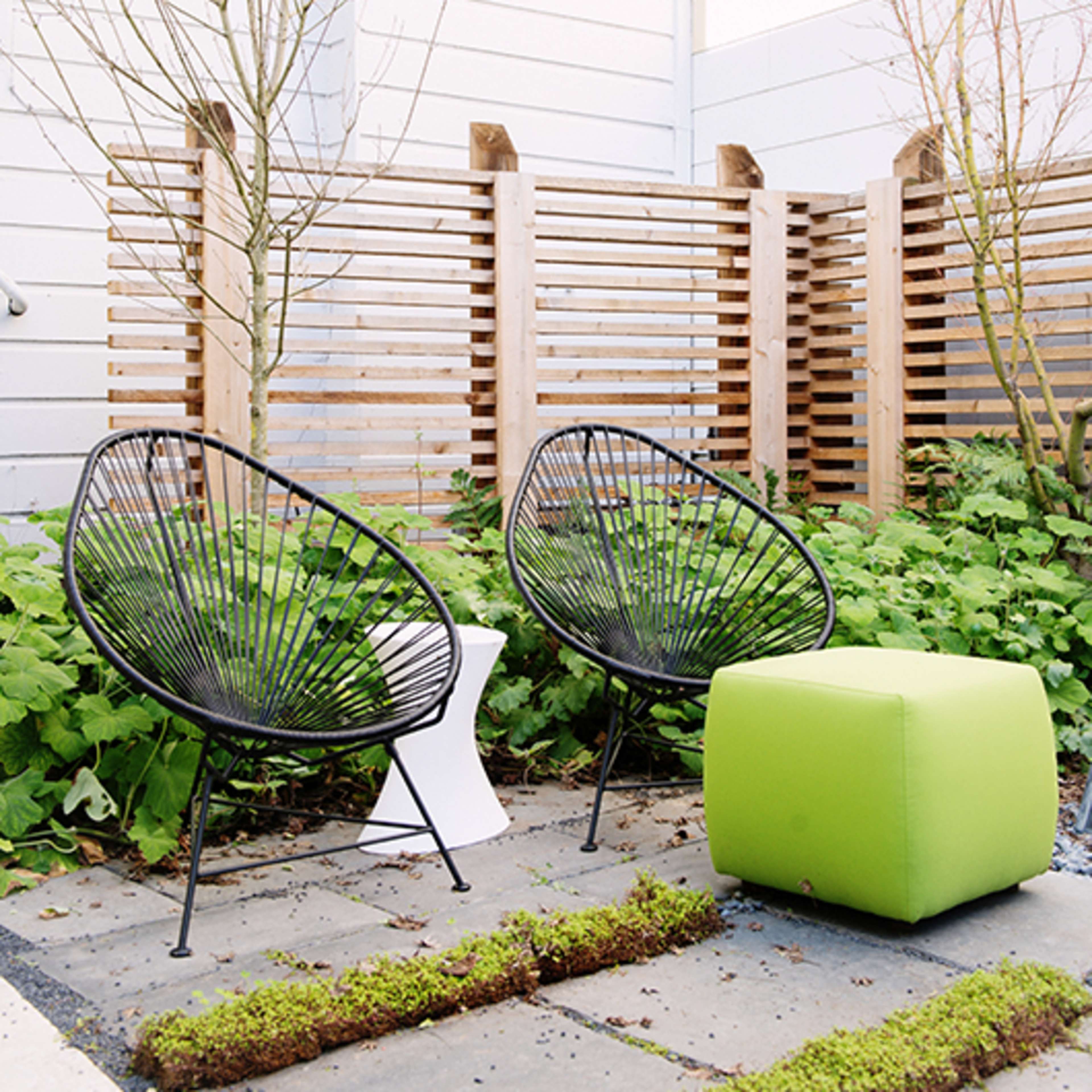 Two black wire chairs and a green ottoman placed on a stone pathway surrounded by greenery and a wooden fence.