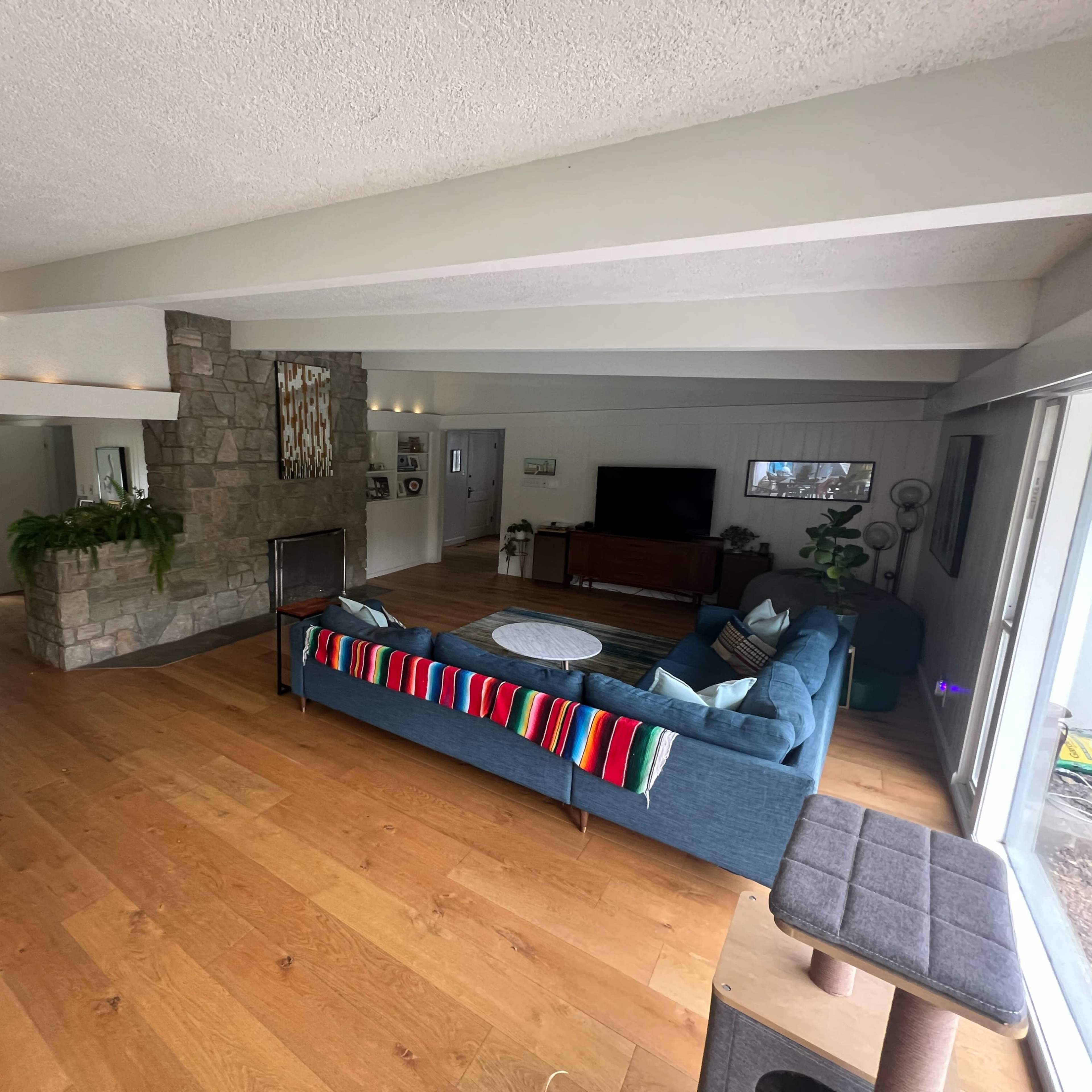 A spacious living room with wooden flooring, a stone fireplace, a blue couch, and large windows providing natural light.