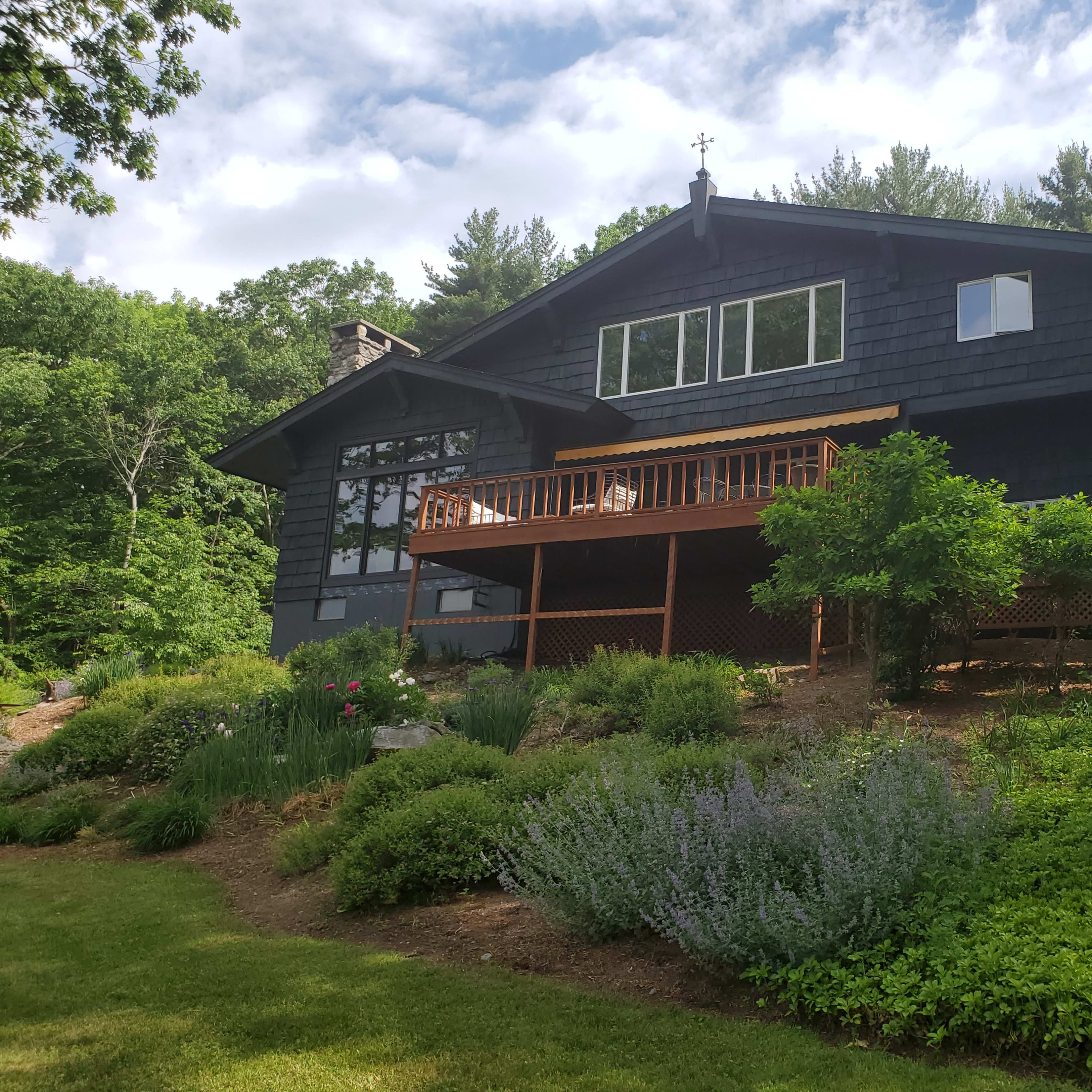 A large dark-colored house with multiple windows and a wooden deck is situated on a landscaped yard featuring various shrubs and flowers.