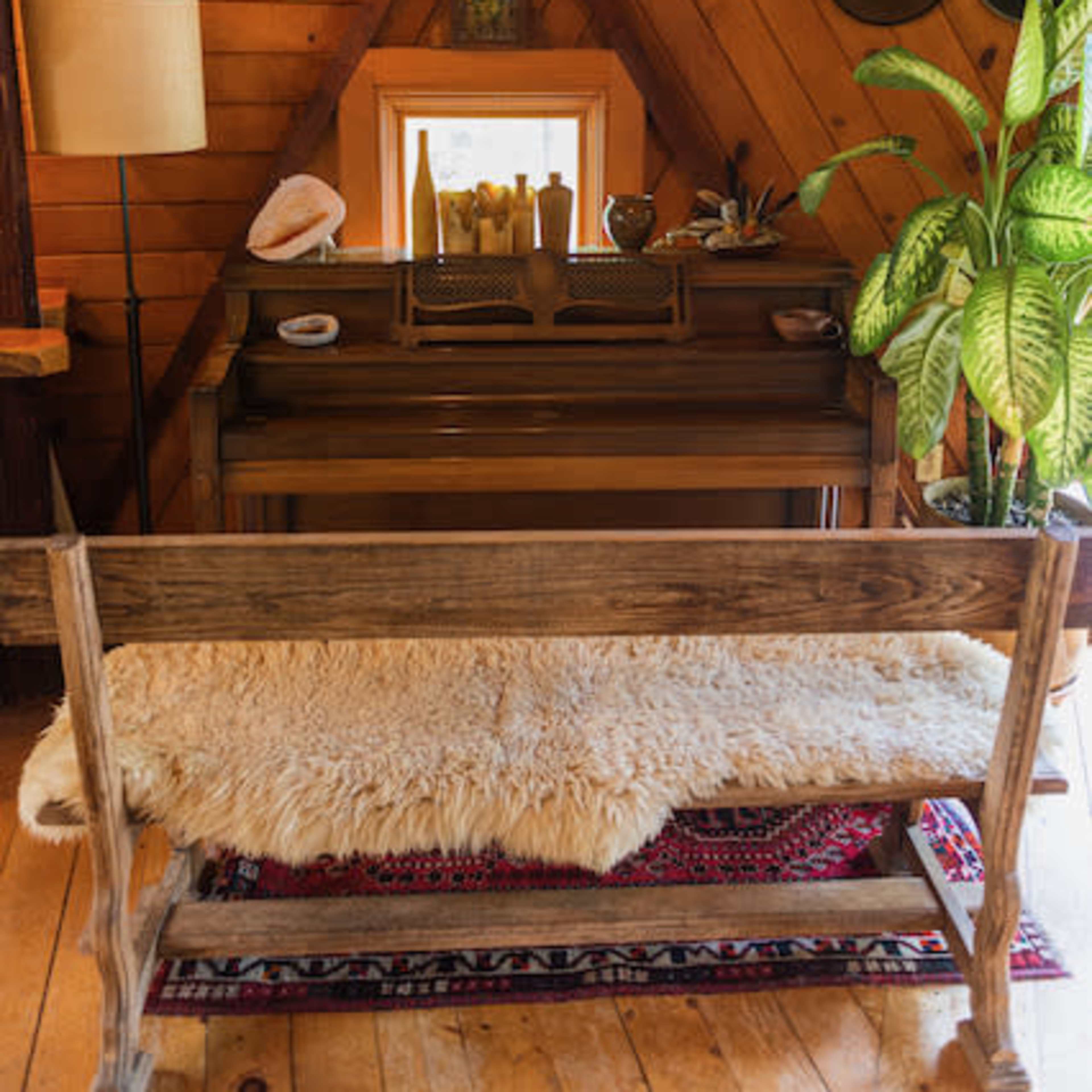 A wooden interior with a piano, a bench covered in a fur rug, and various decorative items, including plants and hats, against wooden walls.
