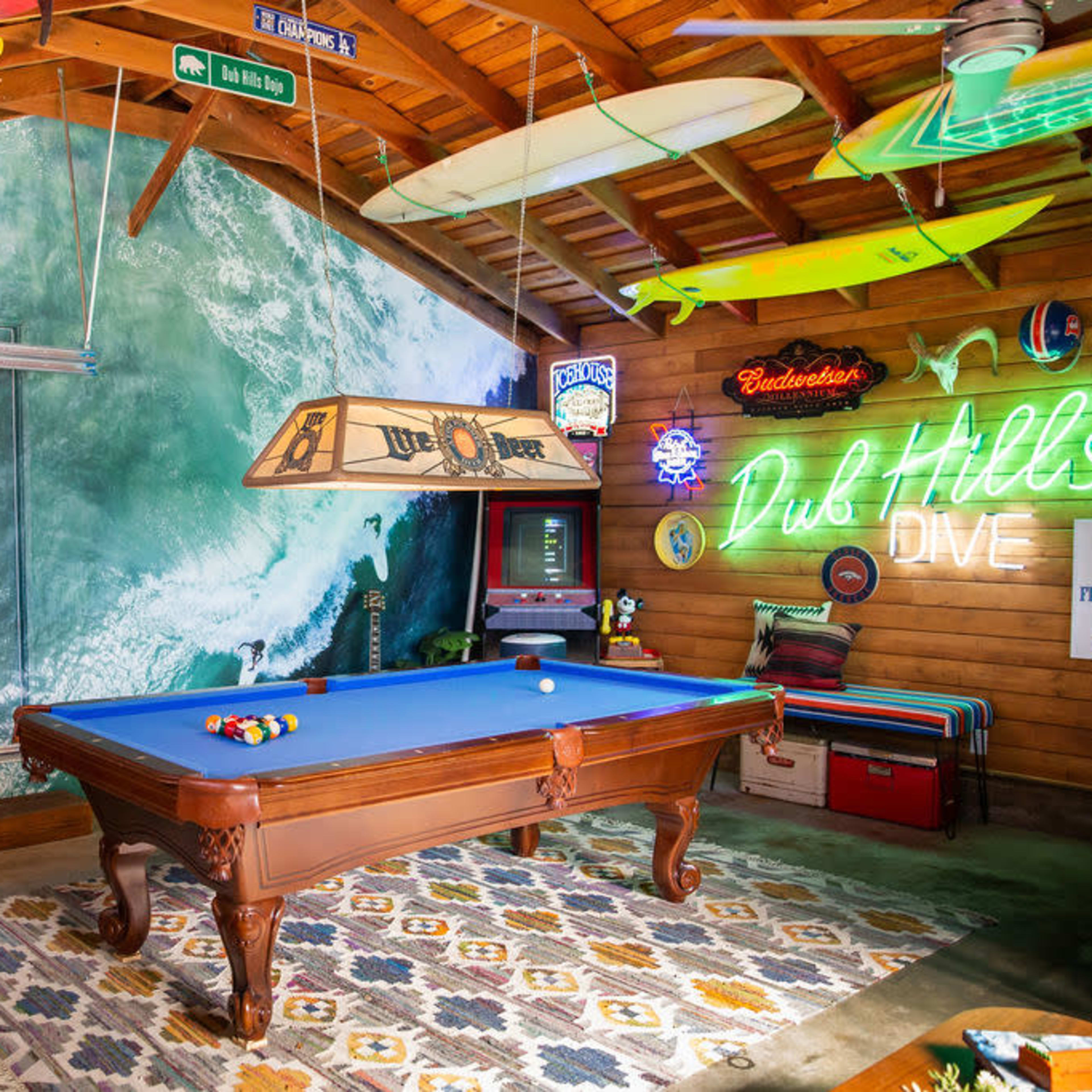 A game room featuring a blue pool table, dartboard, and surf-themed decor, including neon signs and surfboards hanging from the ceiling.