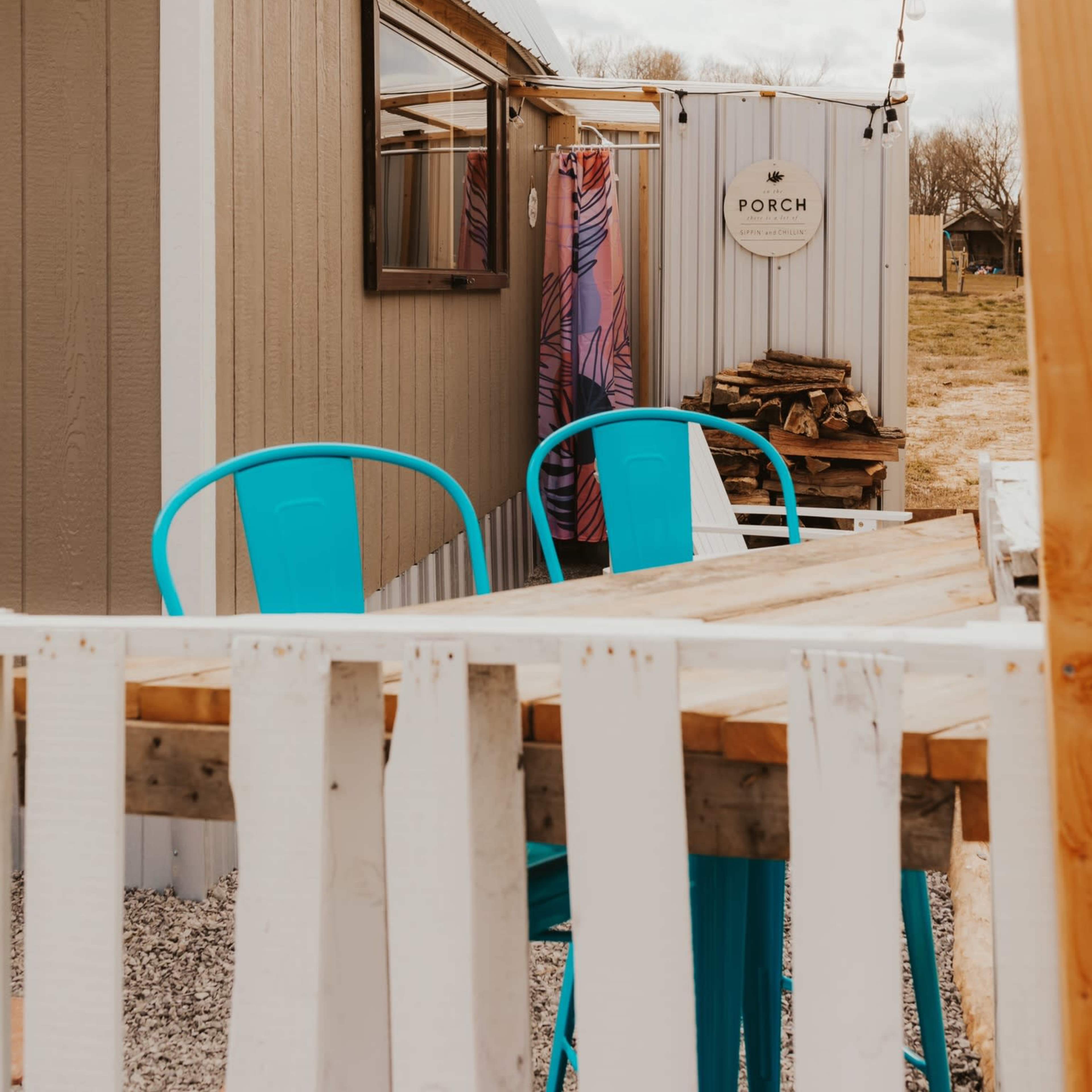 A small cabin with a light-colored exterior features a wooden table and chairs outside, and a nearby shed labeled "PORCH."