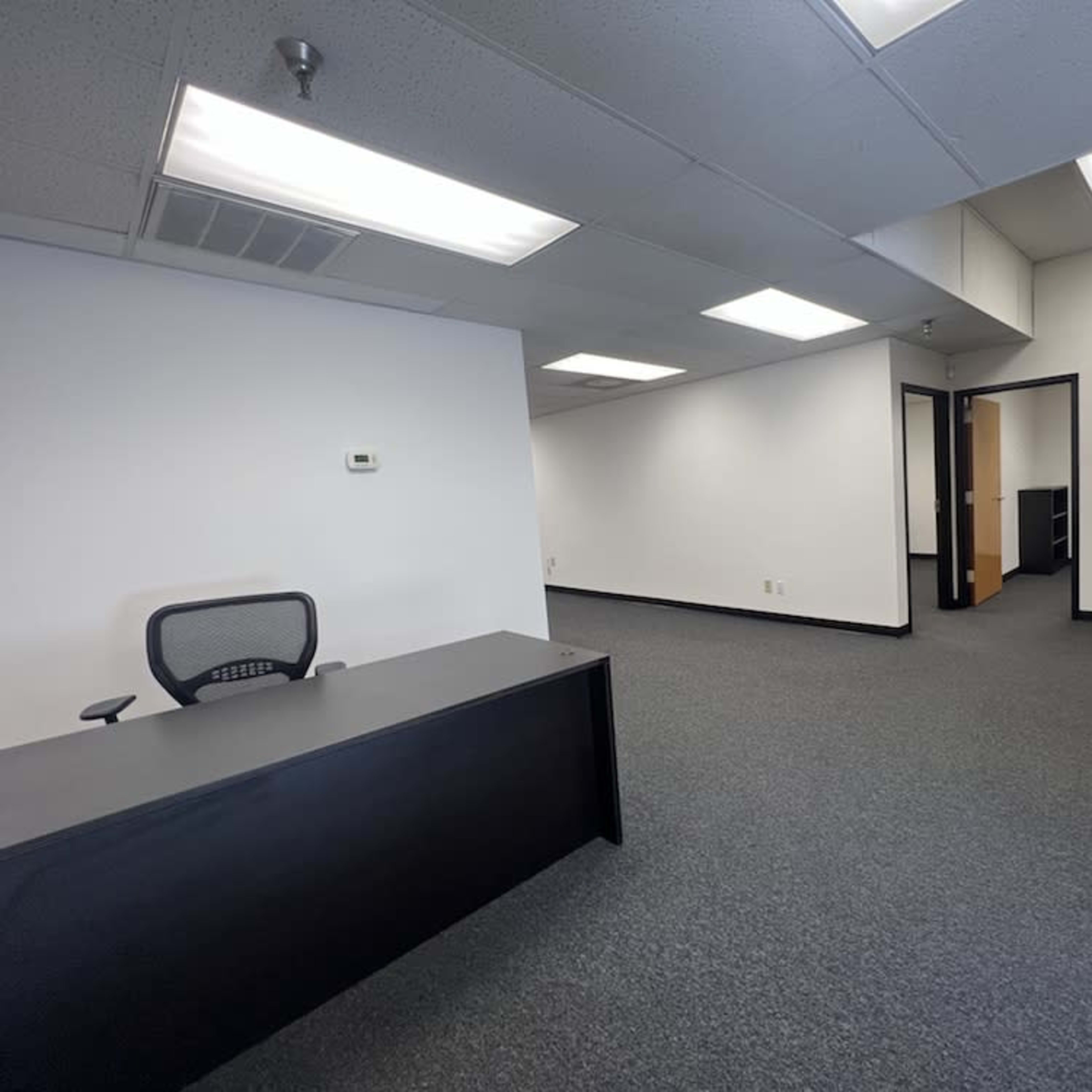 An empty office space with a black reception desk and a waiting area featuring two chairs, all situated on a carpeted floor under simple fluorescent lighting.