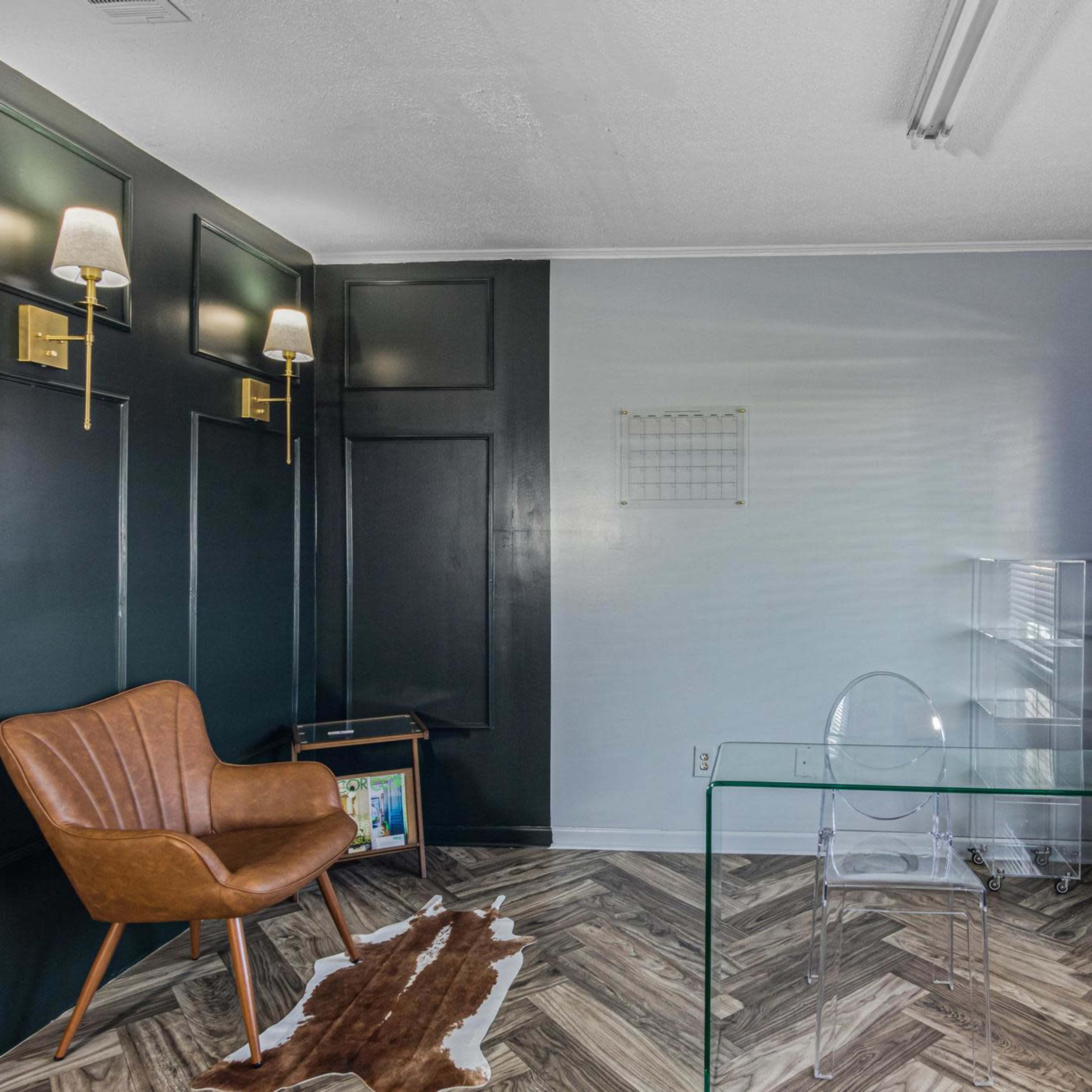 A small office space featuring dark green walls, a hardwood floor arranged in a herringbone pattern, a brown leather chair, a transparent glass-topped table, and a set of blinds covering the window.
