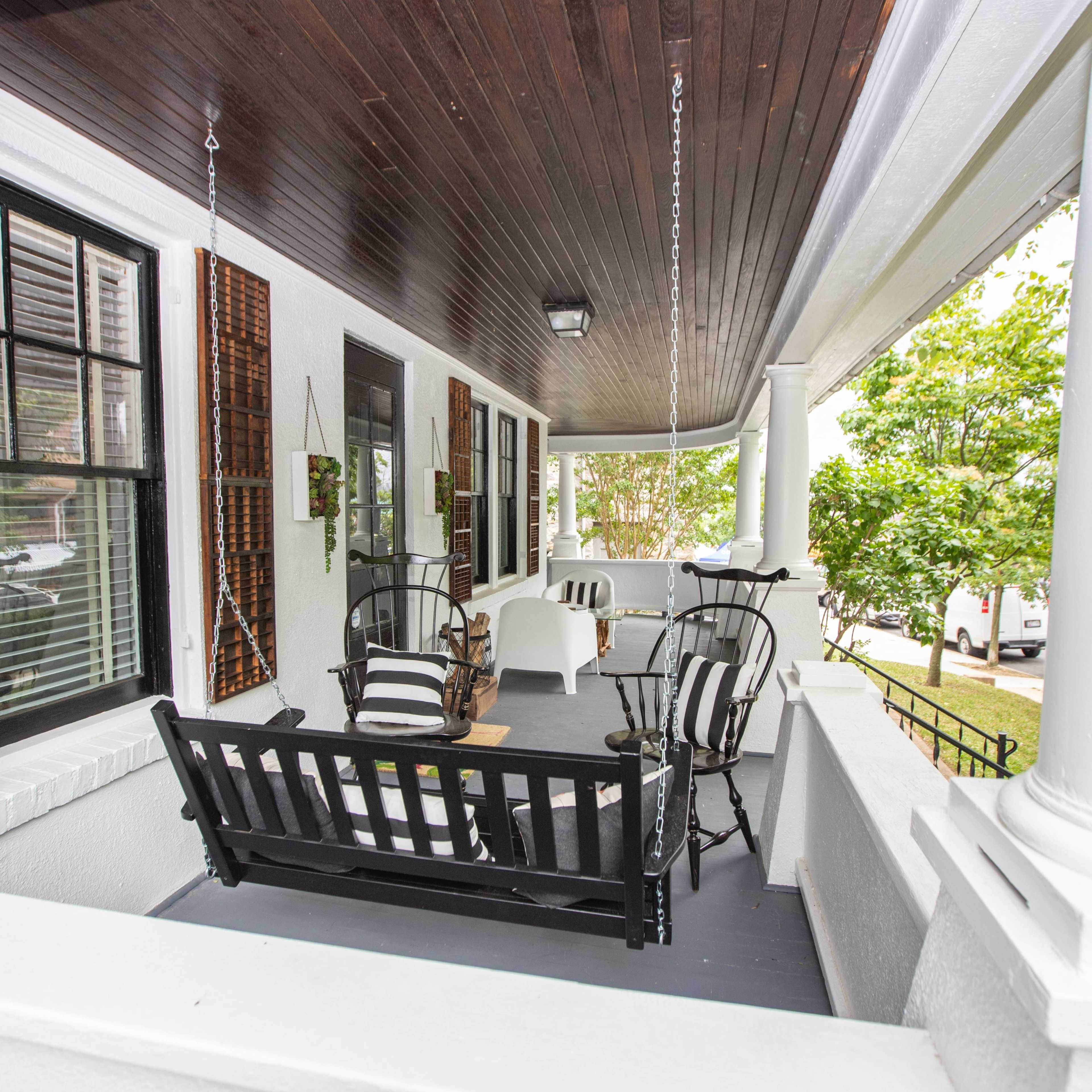 A covered porch featuring black and white furniture, including a swing and chairs, with wooden shutters and columns.