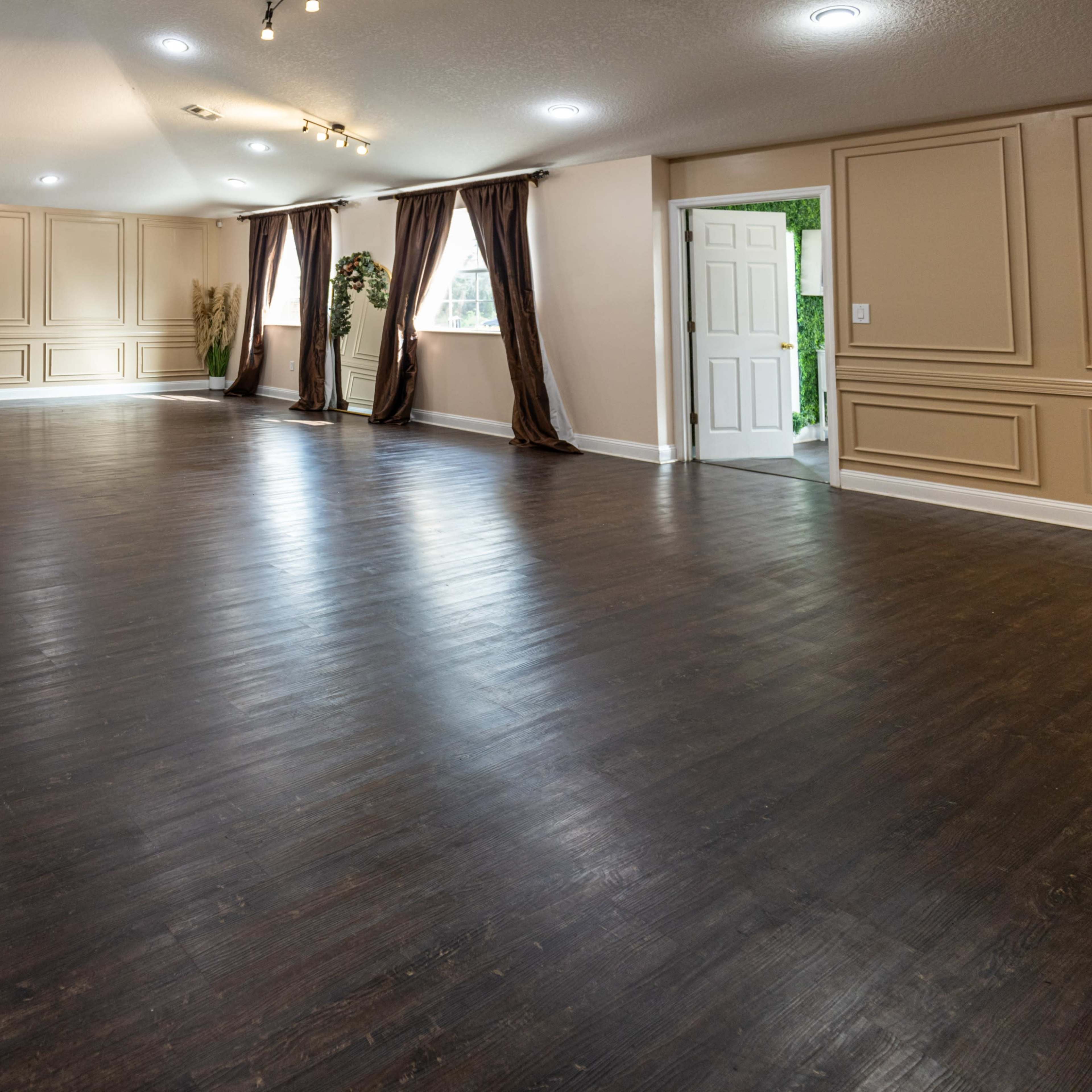 A spacious interior room with dark wood flooring, light-colored walls, and large windows covered by brown curtains.