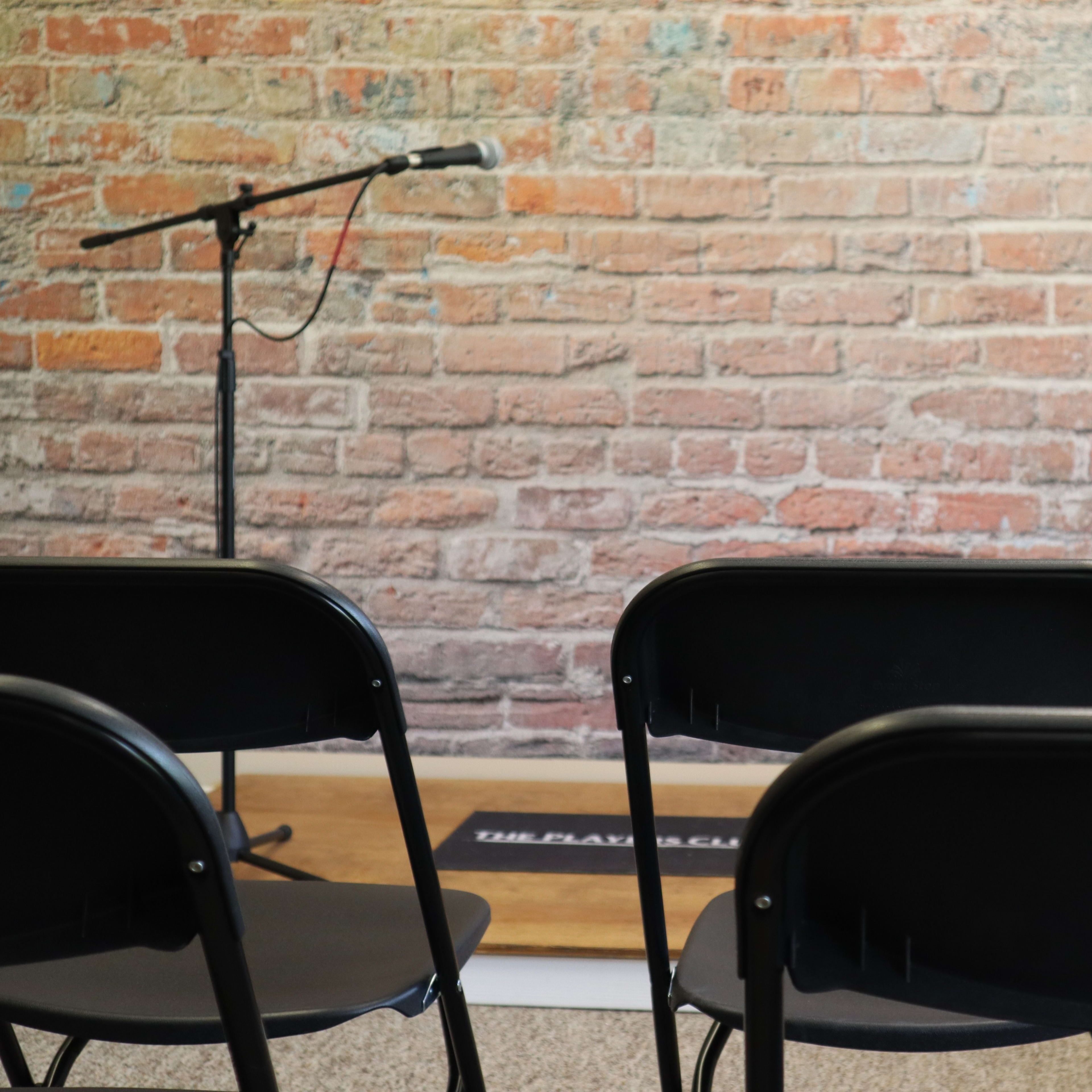 A row of black chairs faces a microphone stand in front of a brick wall with visible texture and color variations.