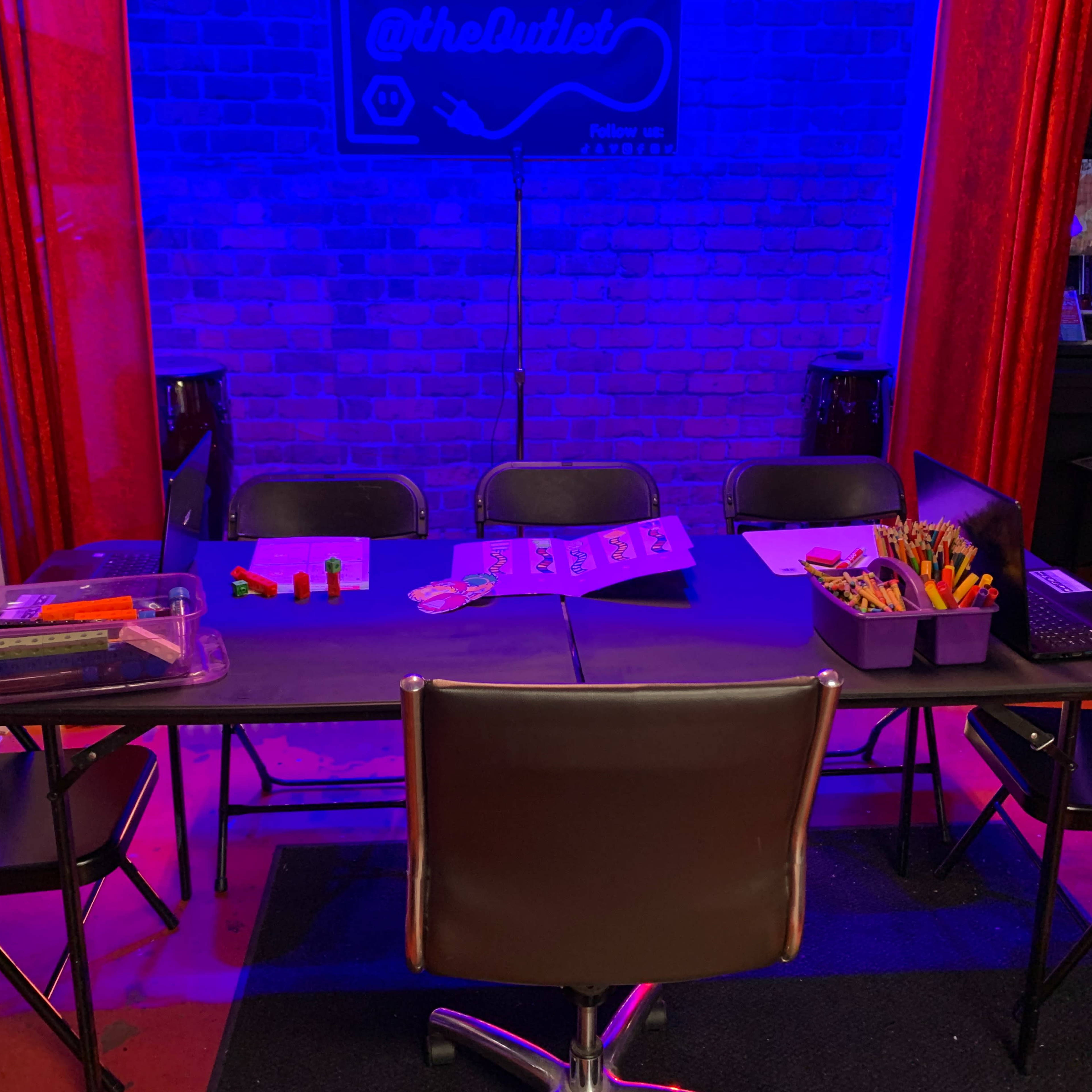A rectangular table with various art supplies and a laptop is set up in a room, surrounded by several black chairs under blue and red lighting.