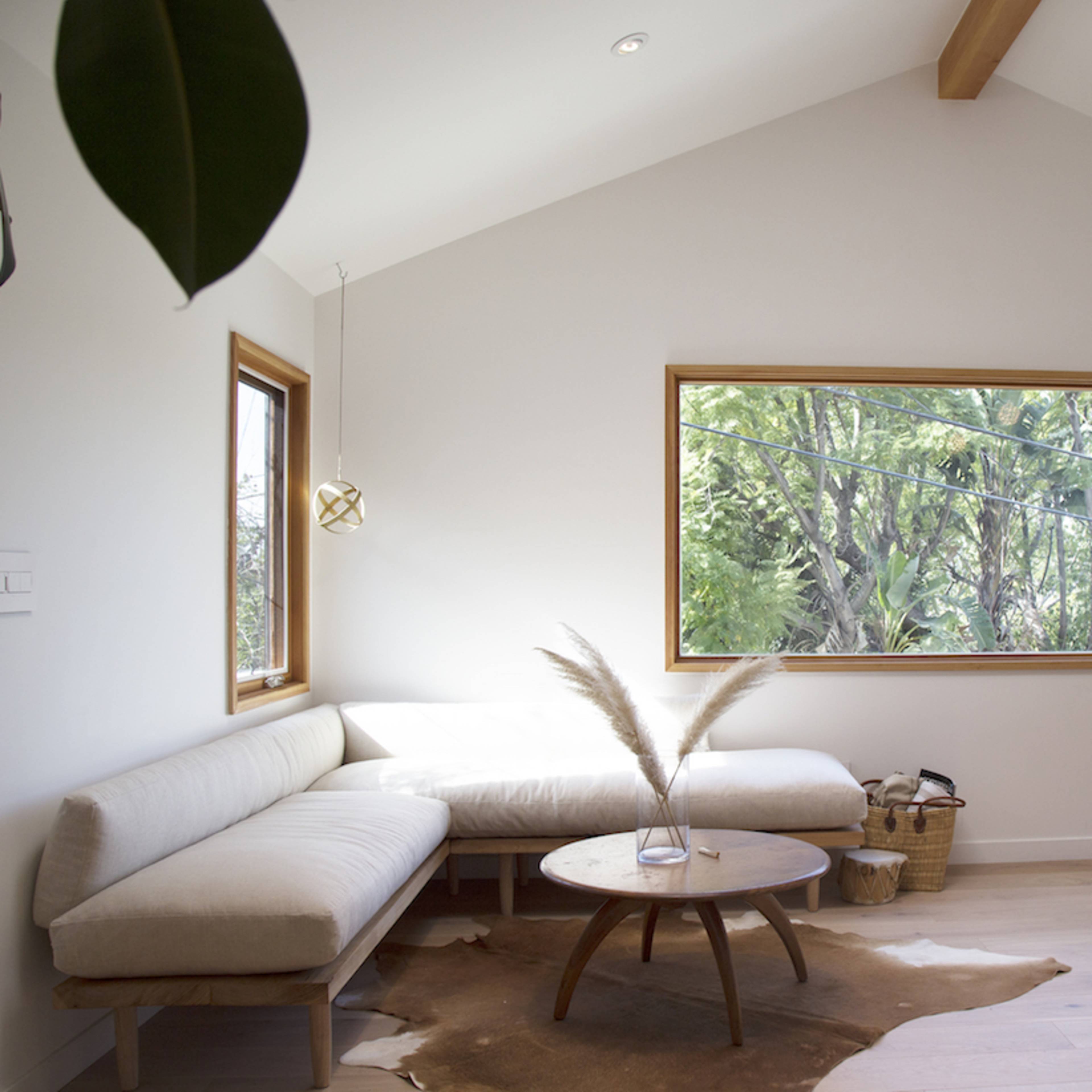 A brightly lit, modern living space featuring a cozy white couch, a round wooden table atop a decorative rug, and large windows overlooking lush greenery.