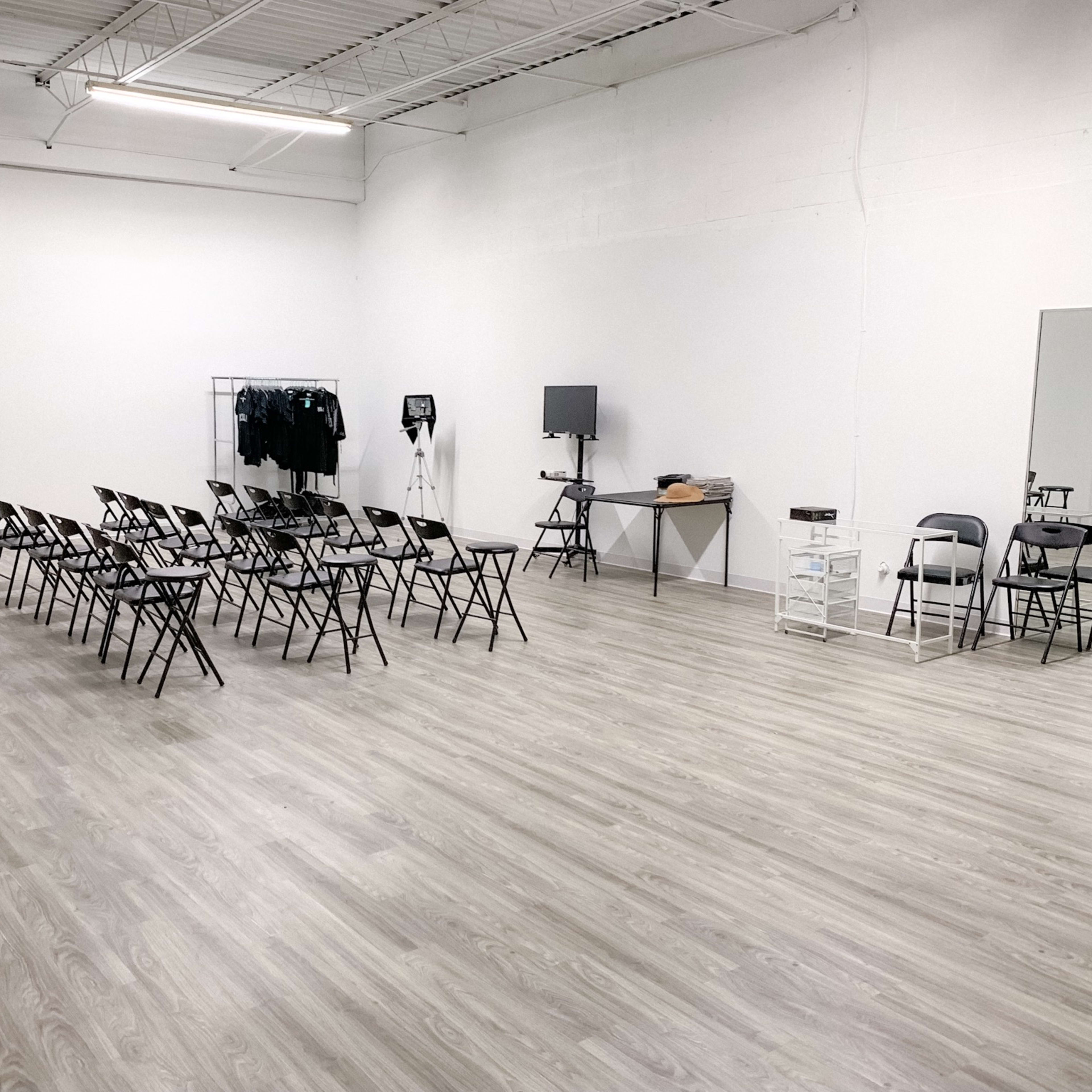 A spacious, empty room with a row of folding chairs facing a wall, a table with equipment, and a large mirror on the opposite side.