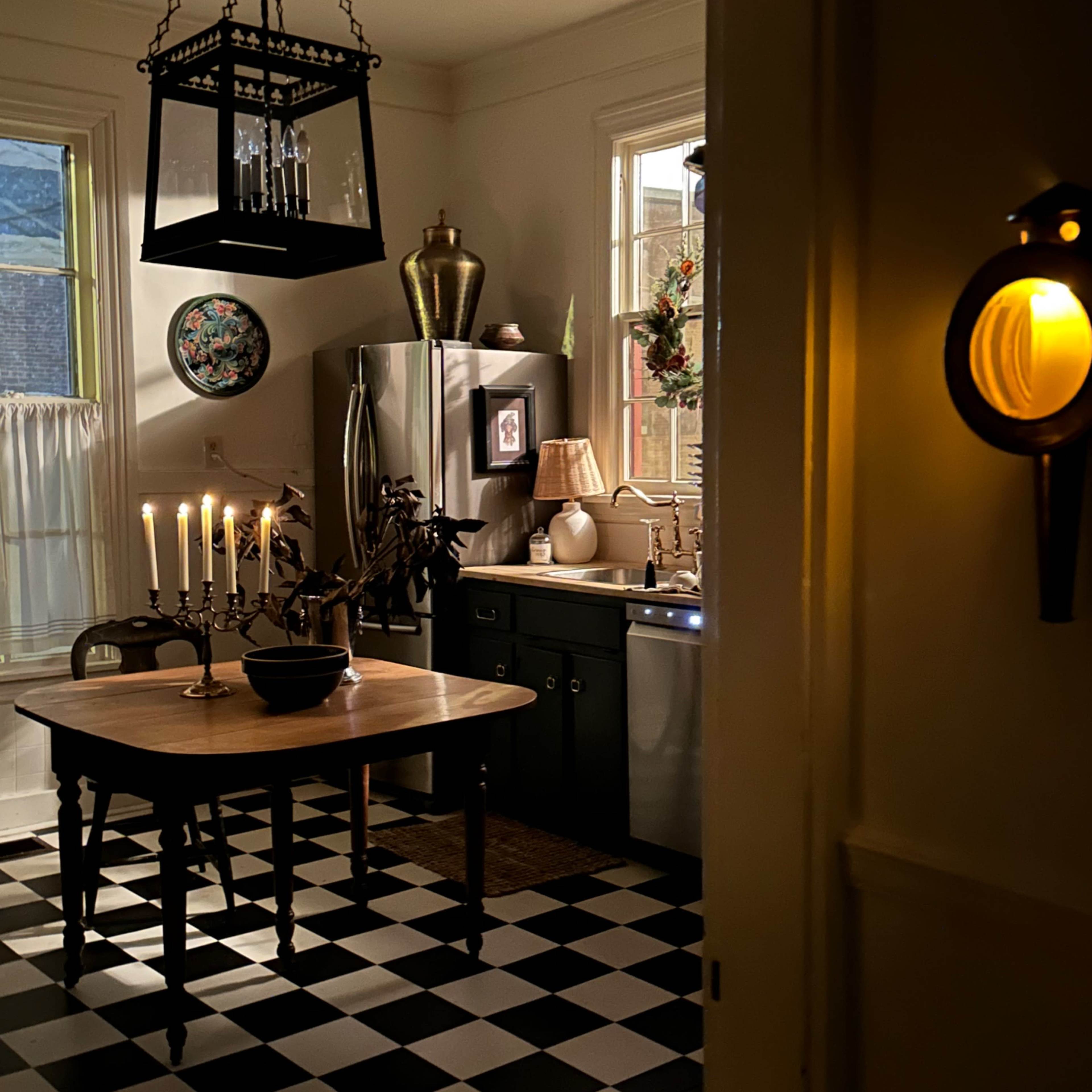 A dimly lit kitchen features a checkered black and white floor, a wooden table, and a chandelier with candles.