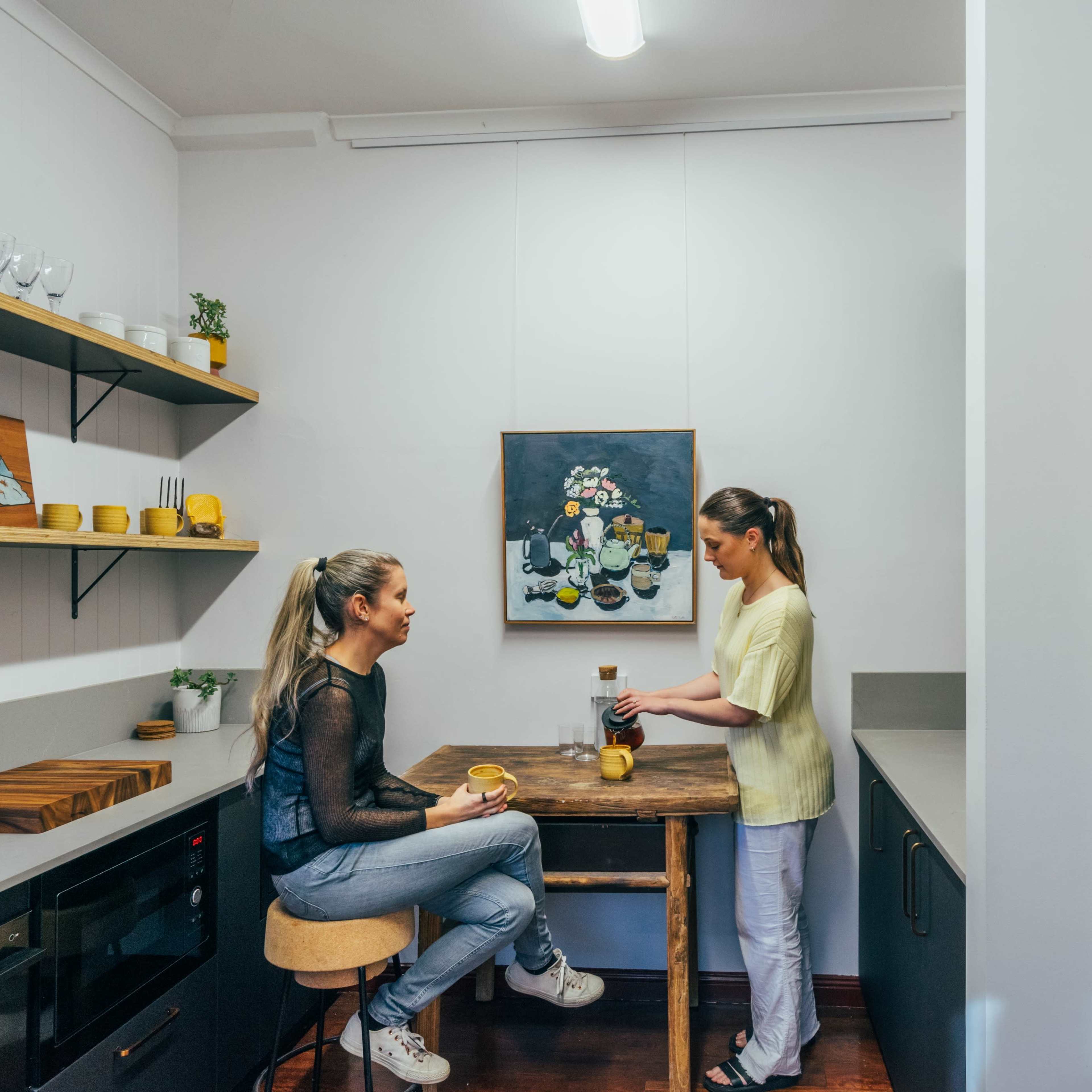 Two women are seated at a wooden table in a modern kitchen while one prepares coffee and the other enjoys a drink.