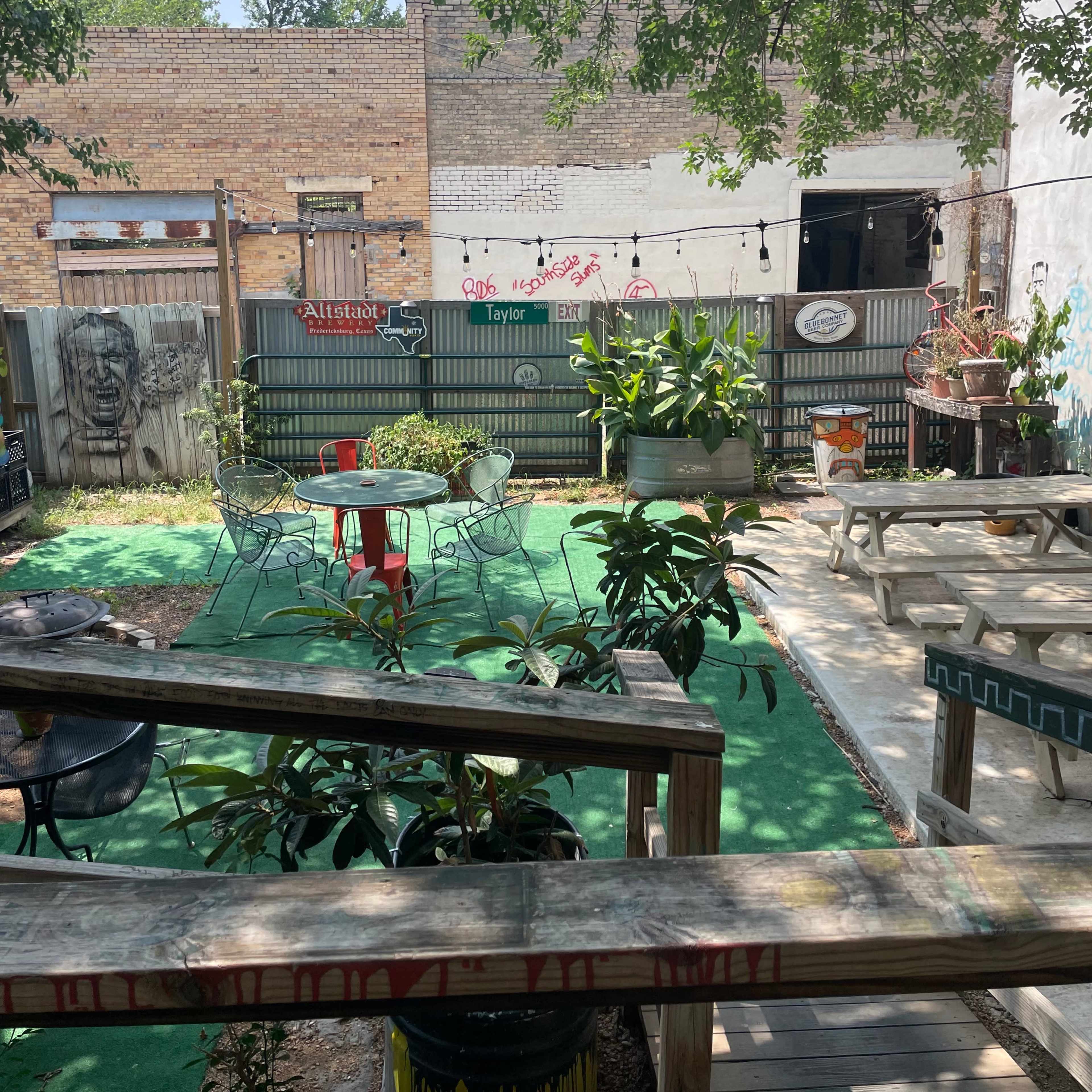 A small outdoor garden area with tables, chairs, plants, and string lights, enclosed by wooden fences and brick walls.
