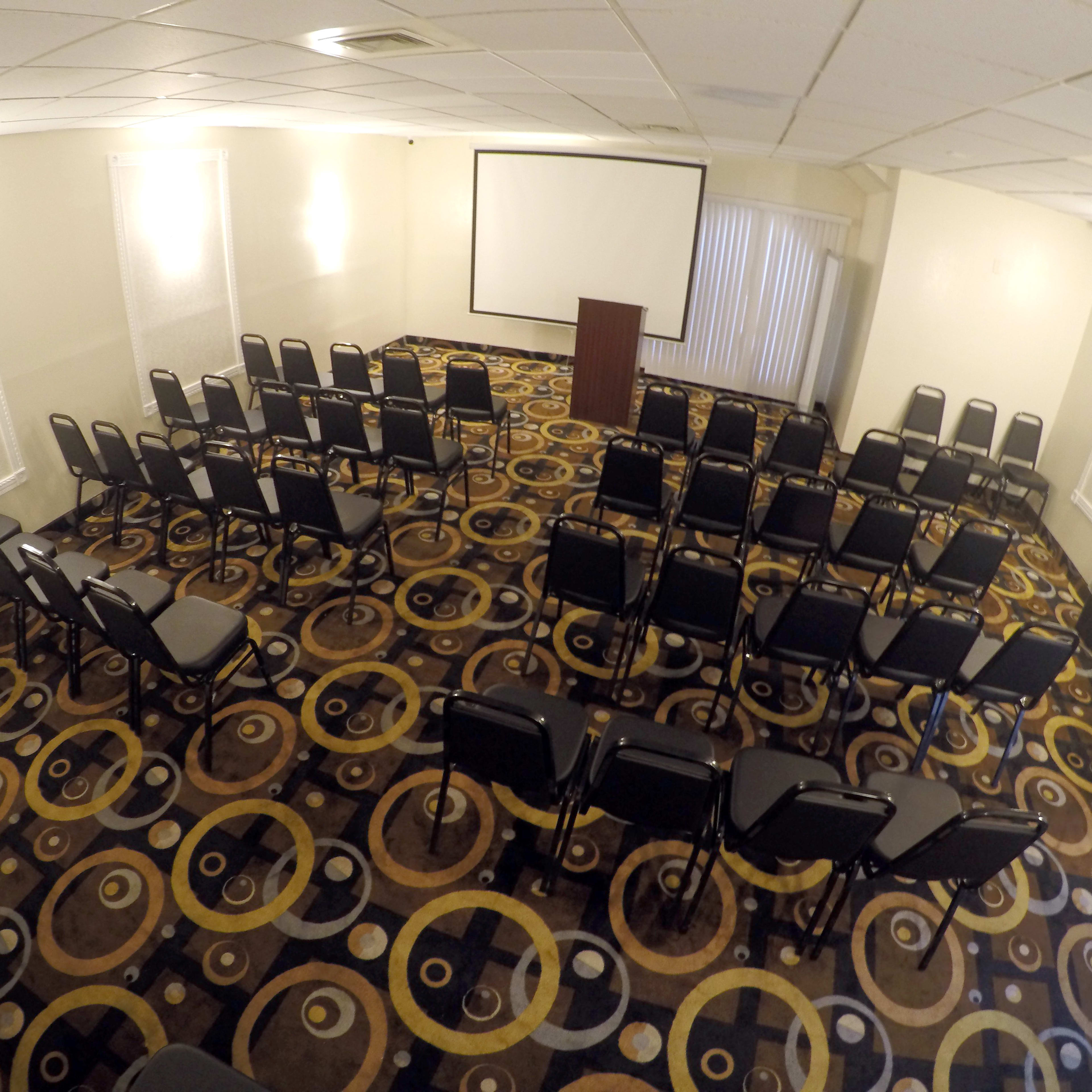 A meeting room is set up with rows of black chairs facing a presentation screen and a podium.