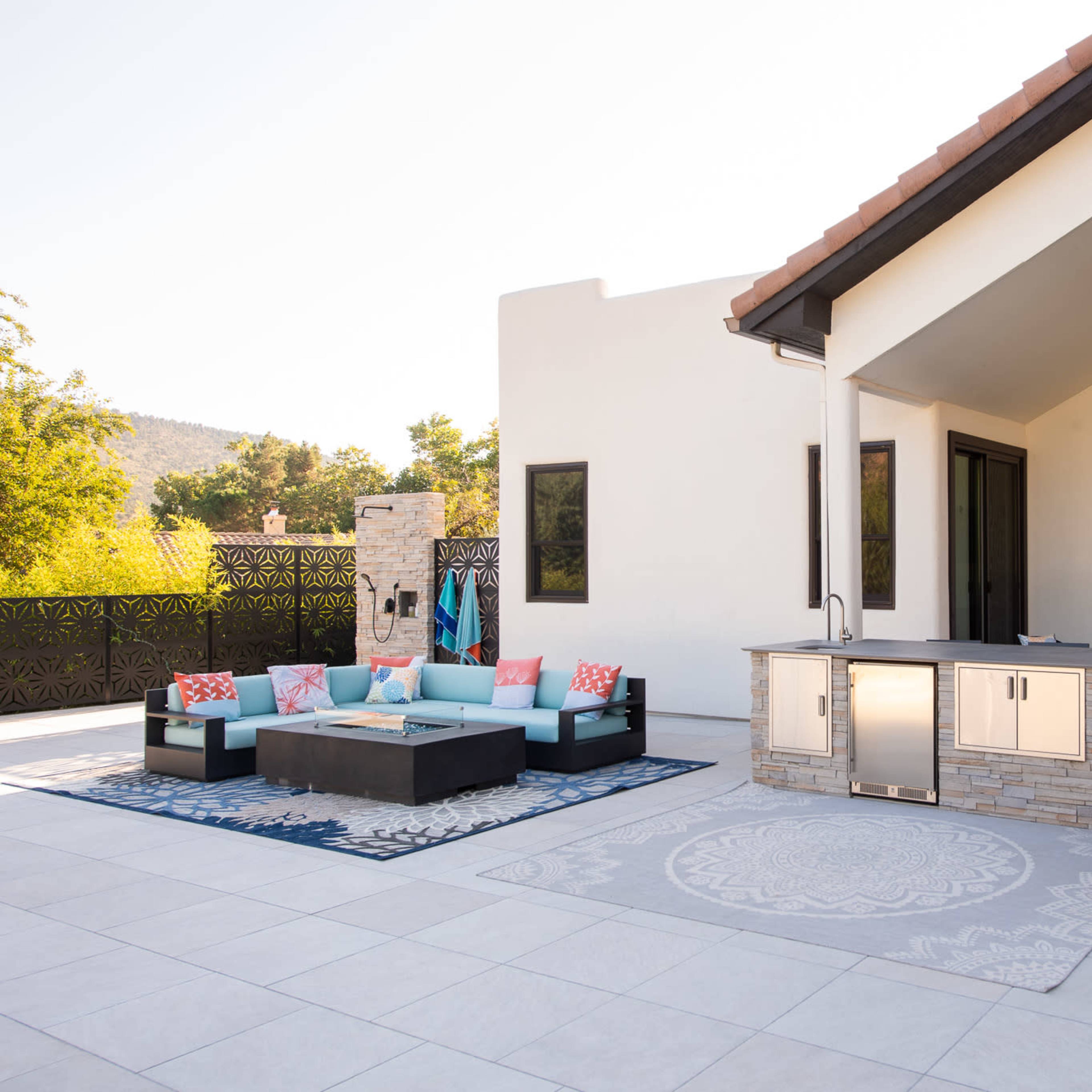 A modern outdoor patio area featuring a structured seating arrangement with a fire pit, an outdoor kitchen, and large windows that open to a view of greenery.