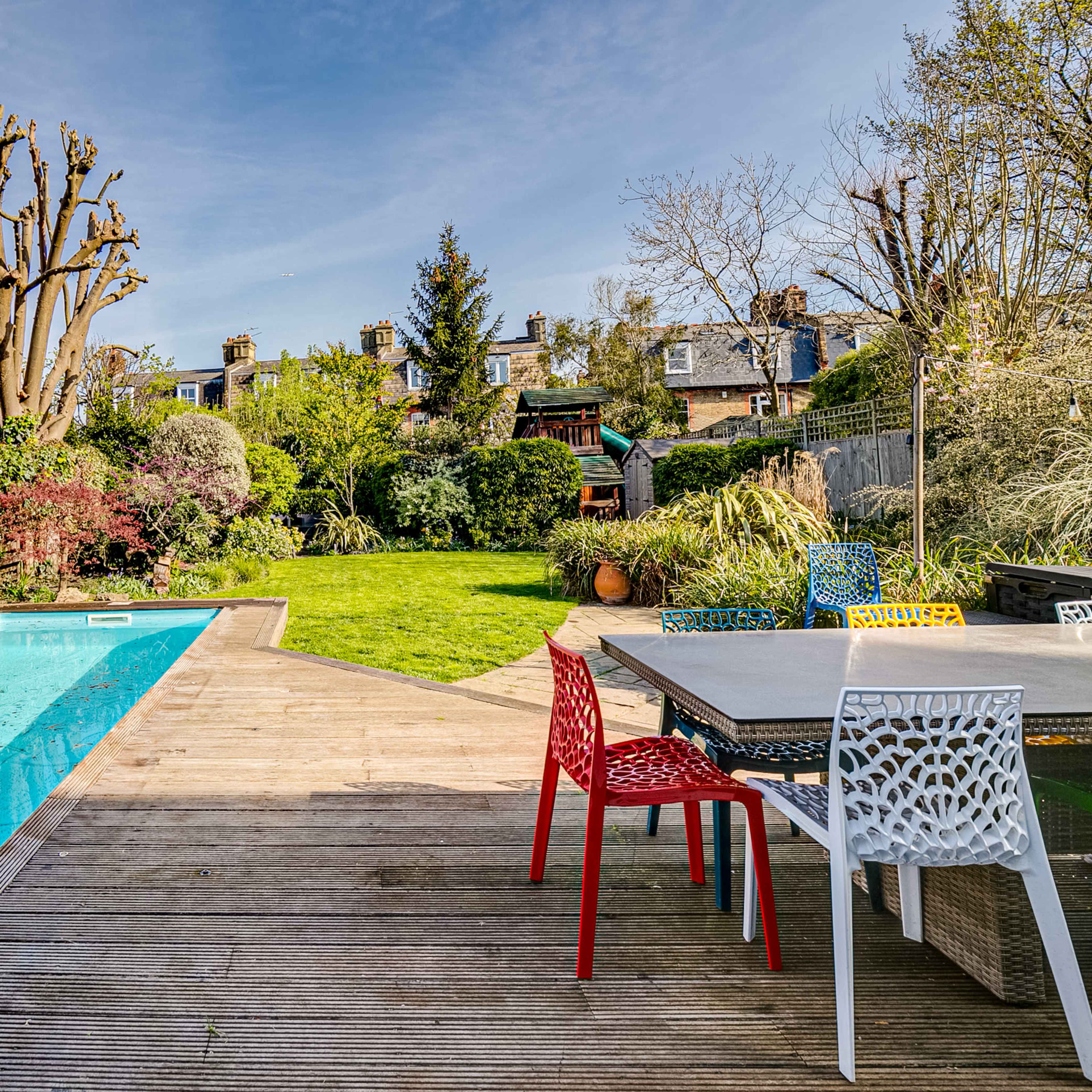 A backyard with a swimming pool, a wooden deck, and a garden filled with greenery and trees.