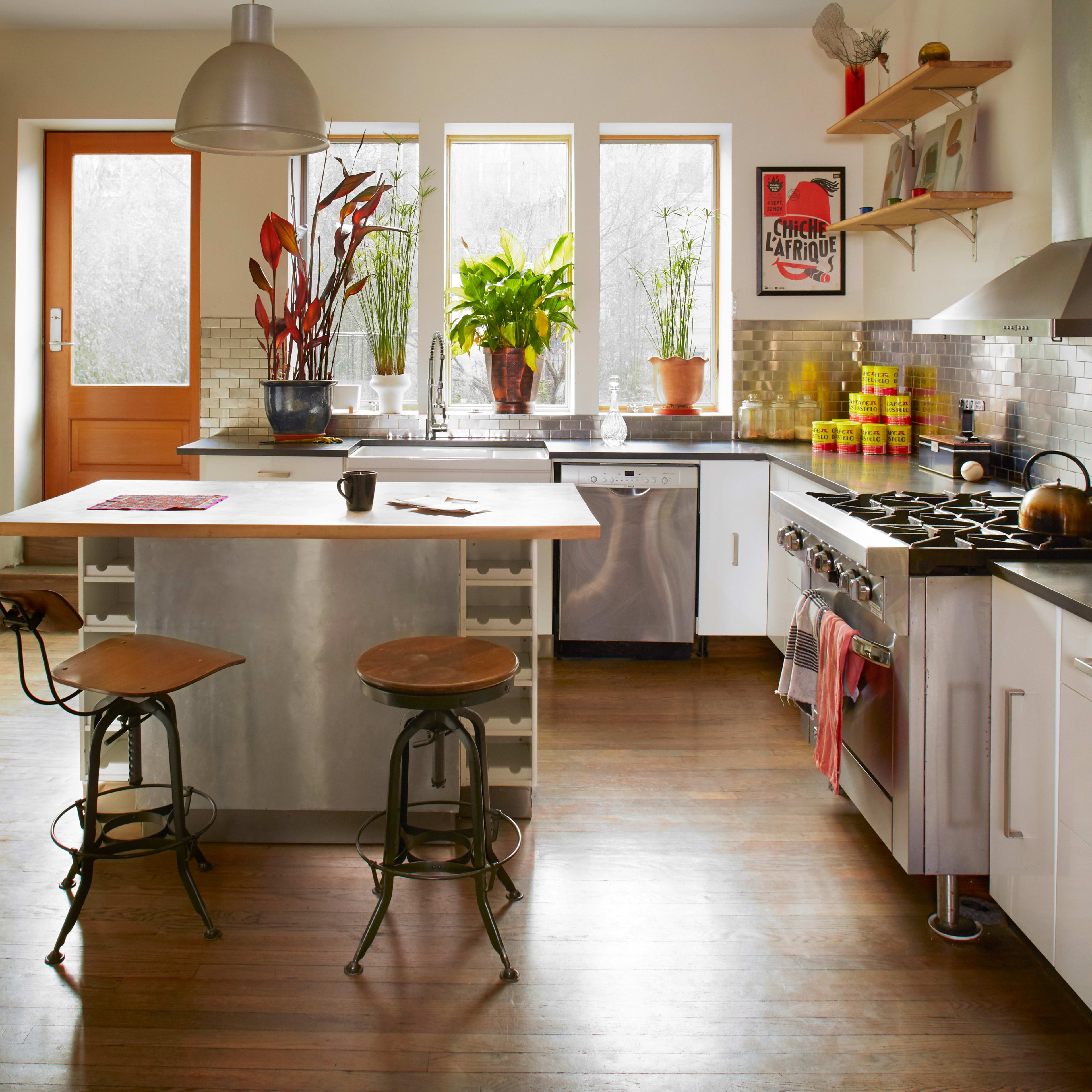 A modern kitchen features a central island with metal seating, stainless steel appliances, and large windows allowing natural light to illuminate the room.