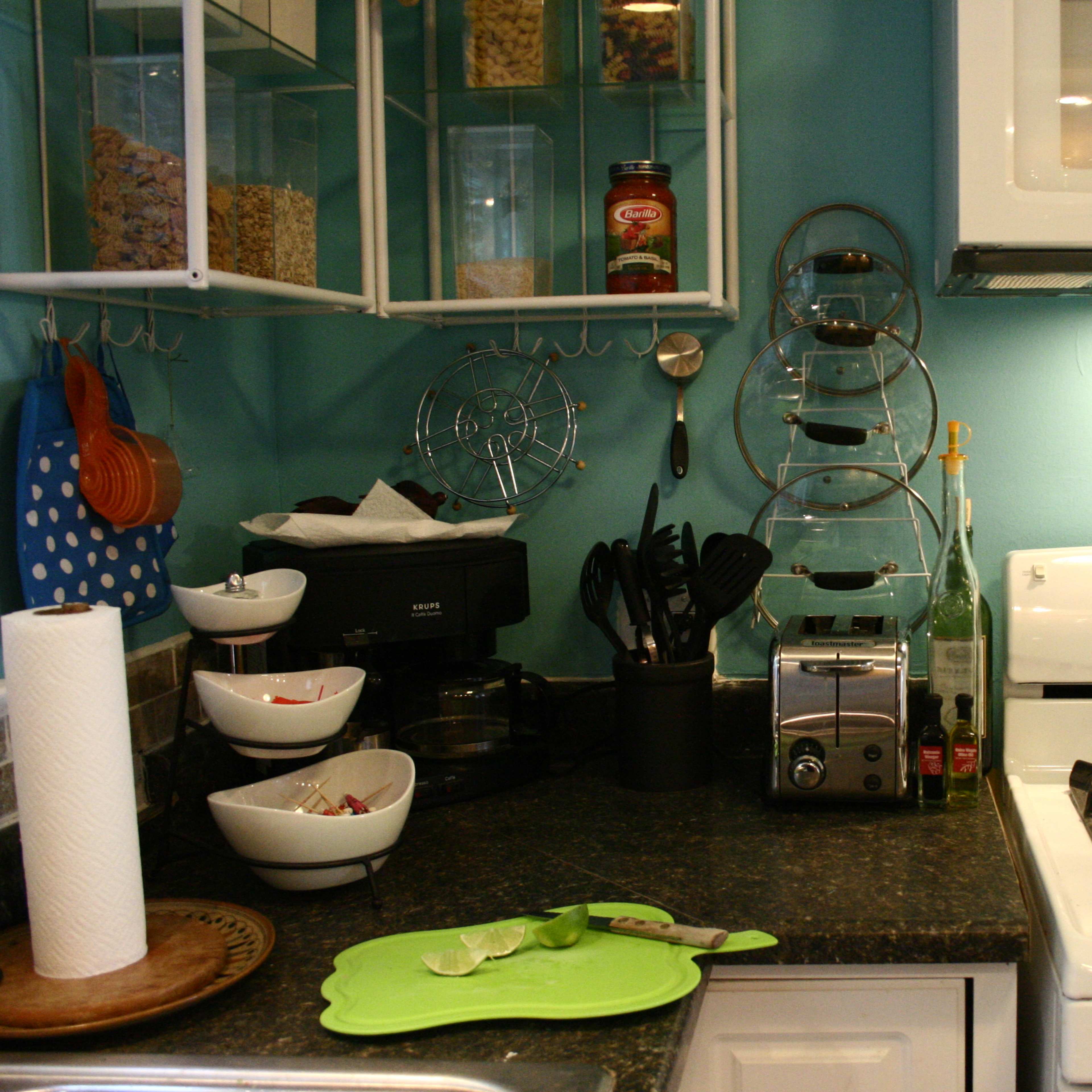 A compact kitchen features a gas stove, a toaster, various utensils organized in containers, and bowls on a dark countertop, with a teal wall as the backdrop.
