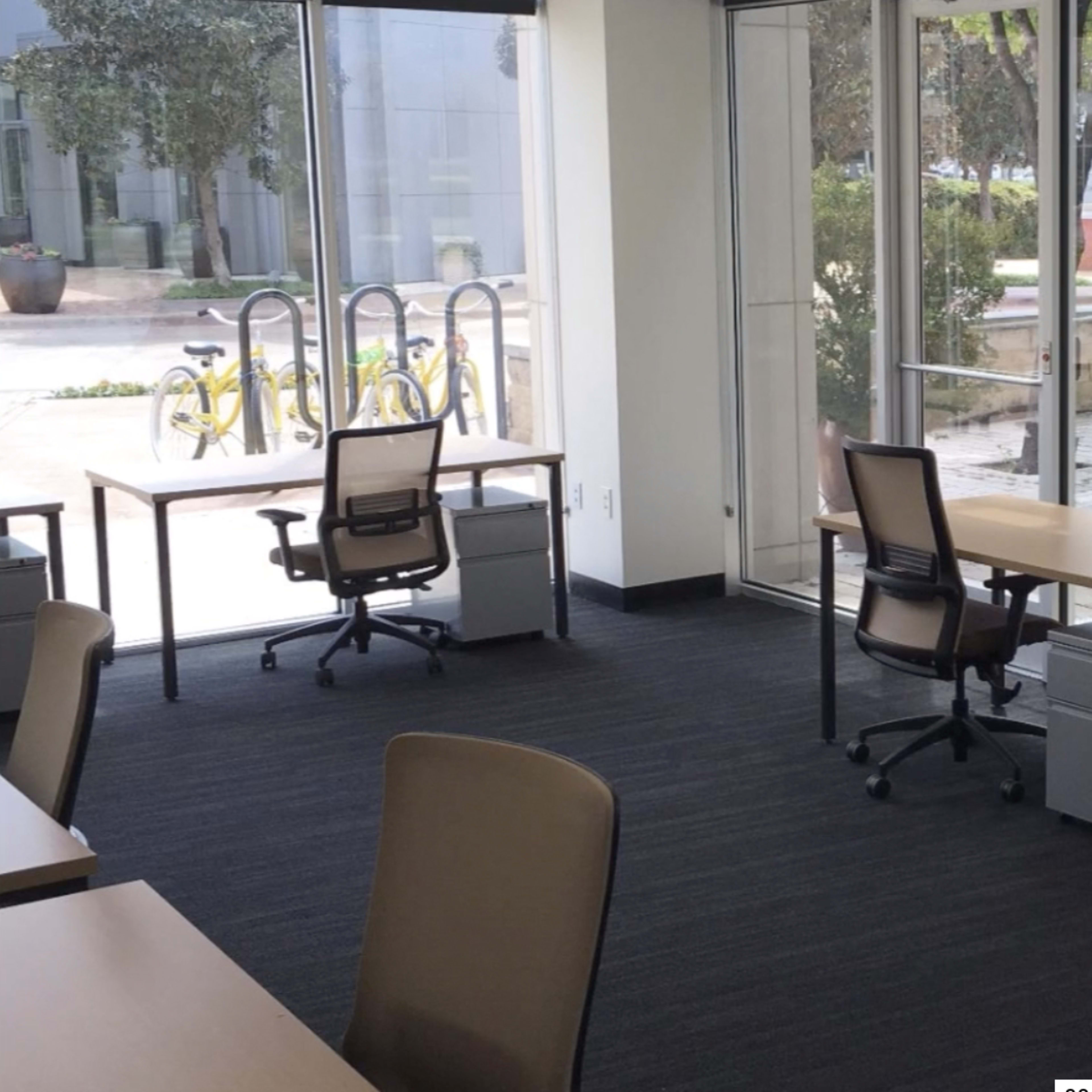 A modern office space with two desks and chairs beside large windows overlooking a courtyard.