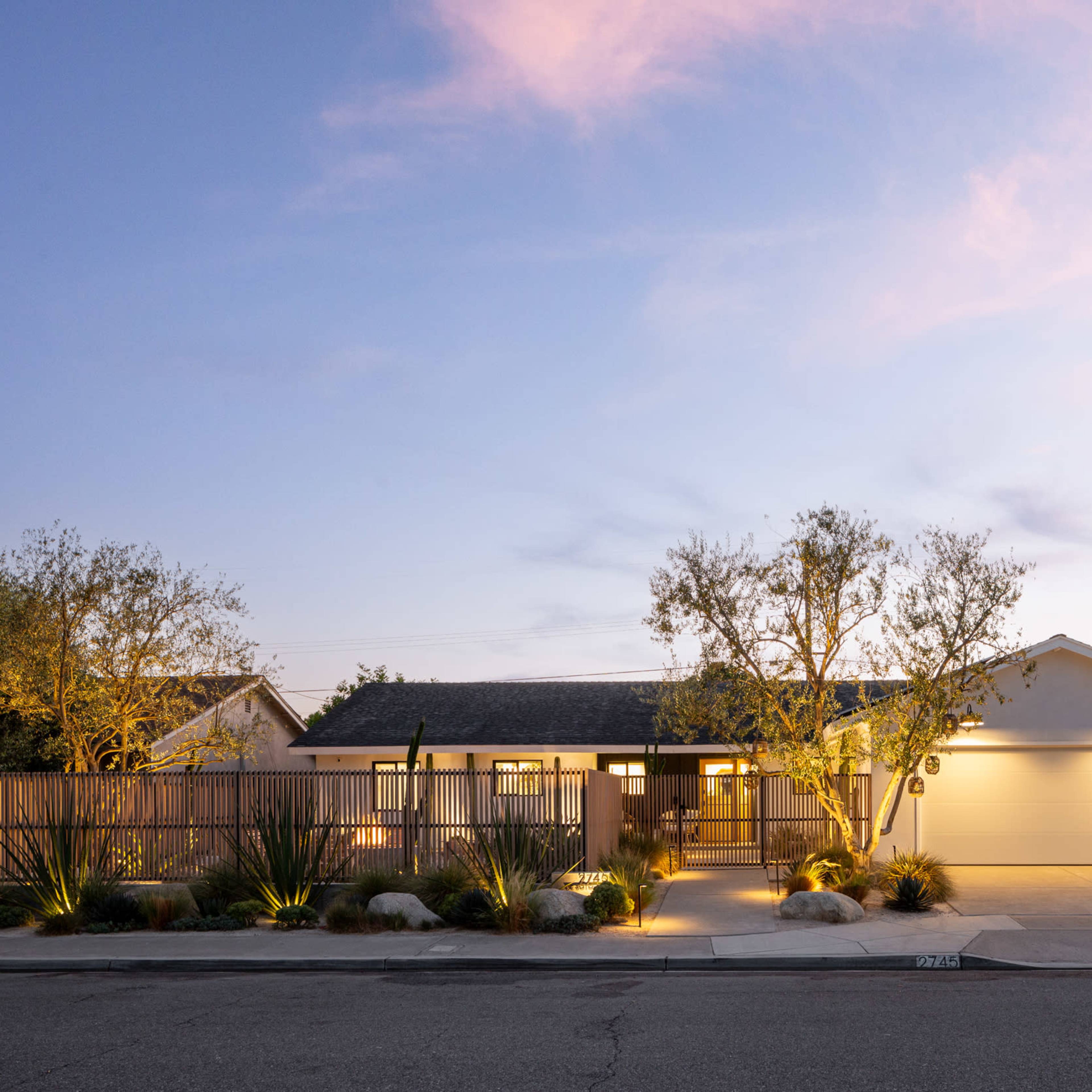 A contemporary single-story house with a landscaped front yard is illuminated under a twilight sky.