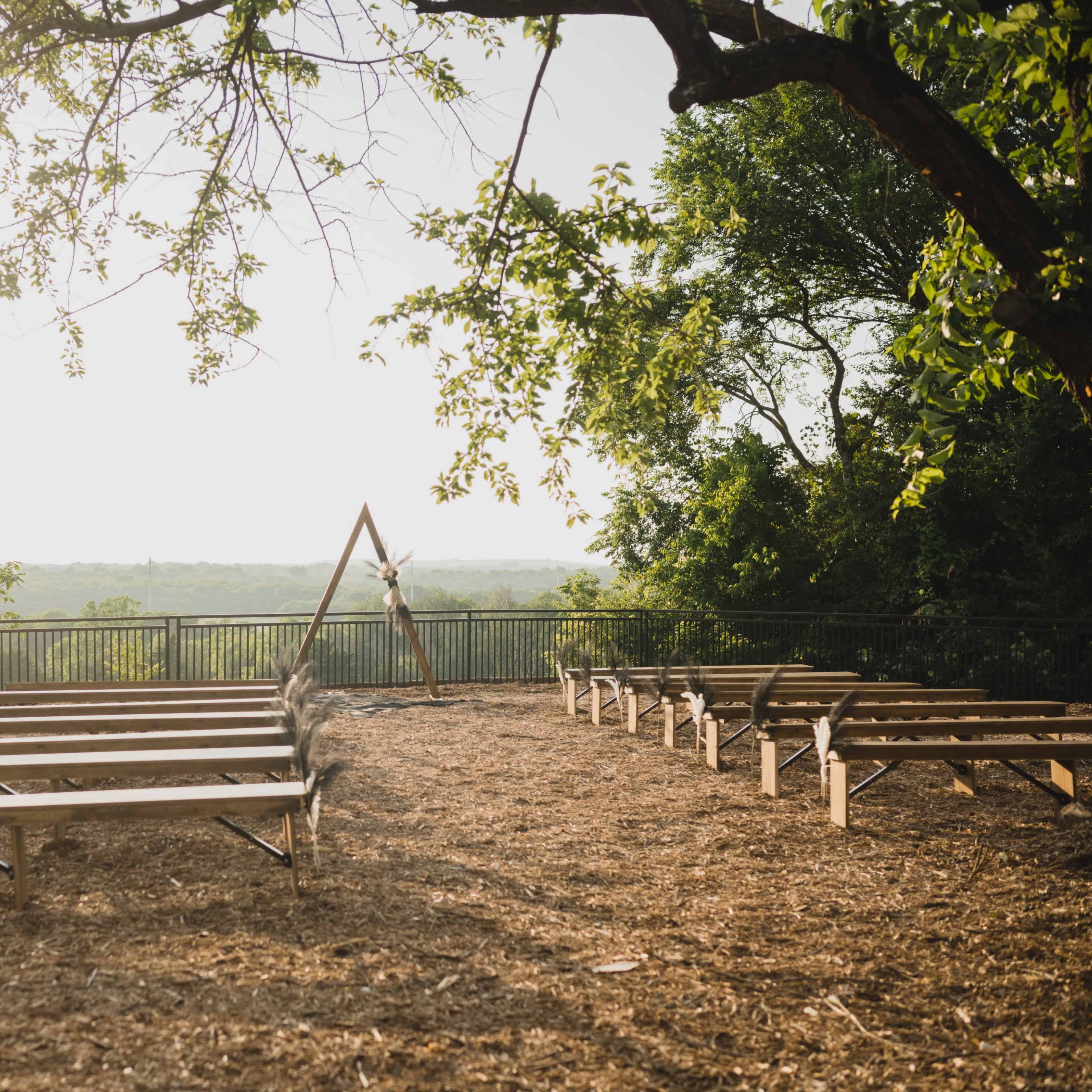 A rustic outdoor venue with wooden benches arranged in a semicircle facing an arched structure, surrounded by trees and overlooking a scenic view.