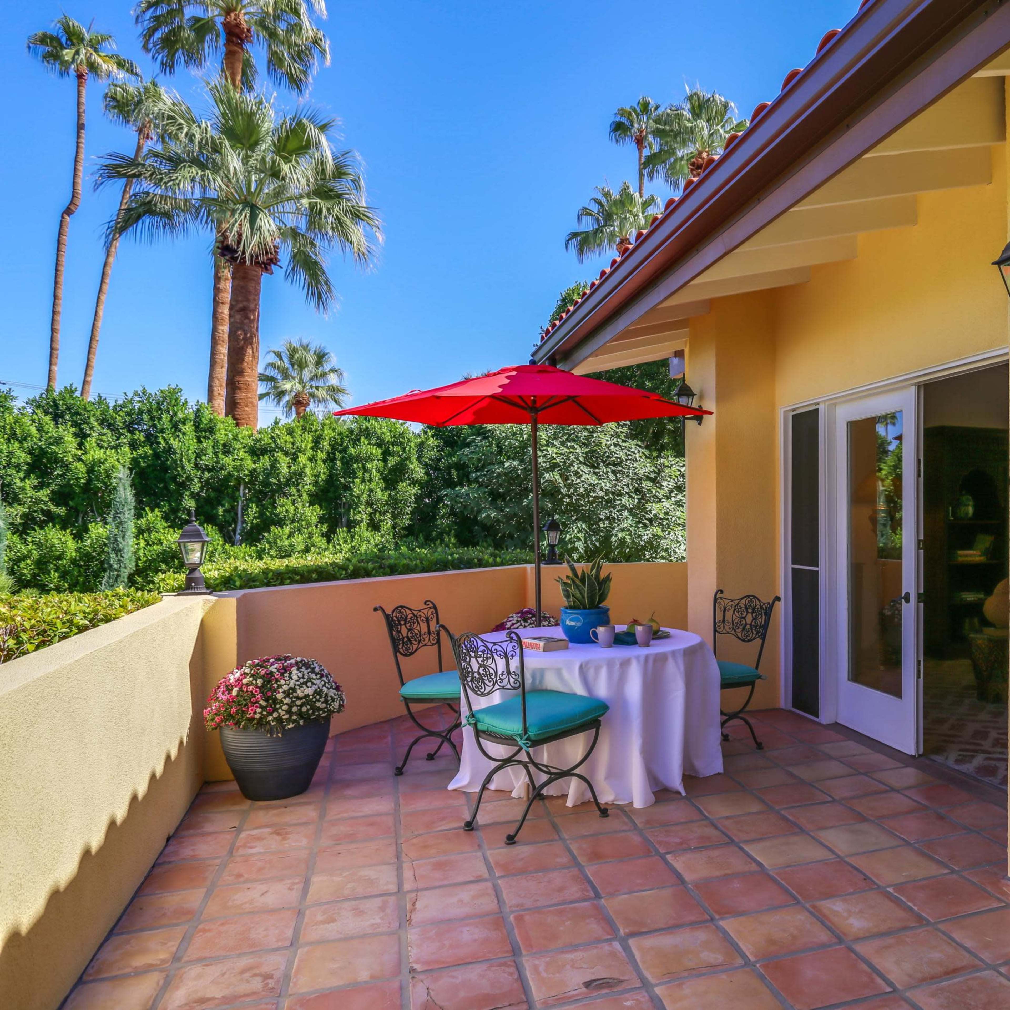 A patio features a table with an umbrella, surrounded by potted plants and candle holders against a backdrop of palm trees.