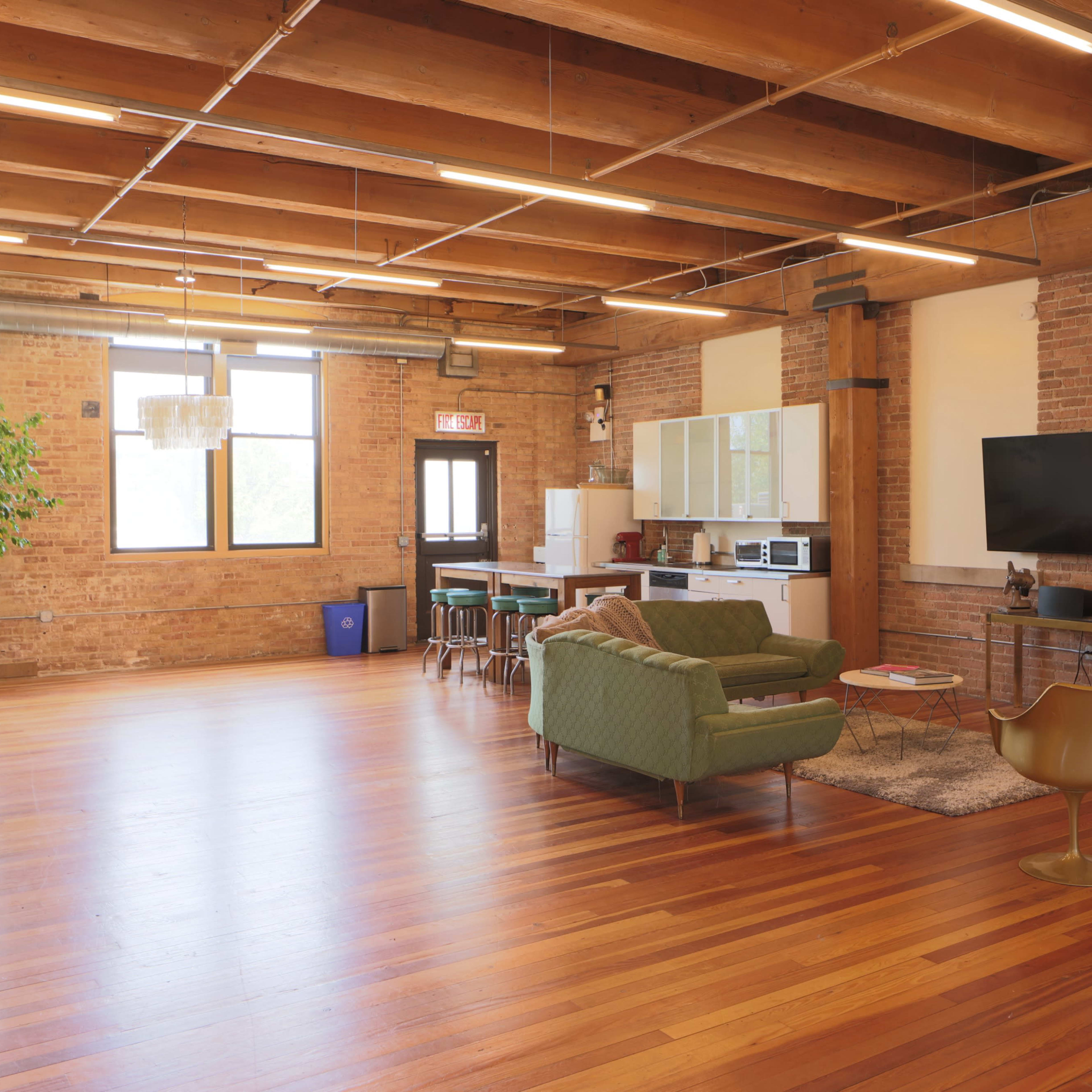 A spacious, modern loft with wooden beams, large windows, a living area featuring a green couch and two gold chairs, and a kitchen area against a brick wall.