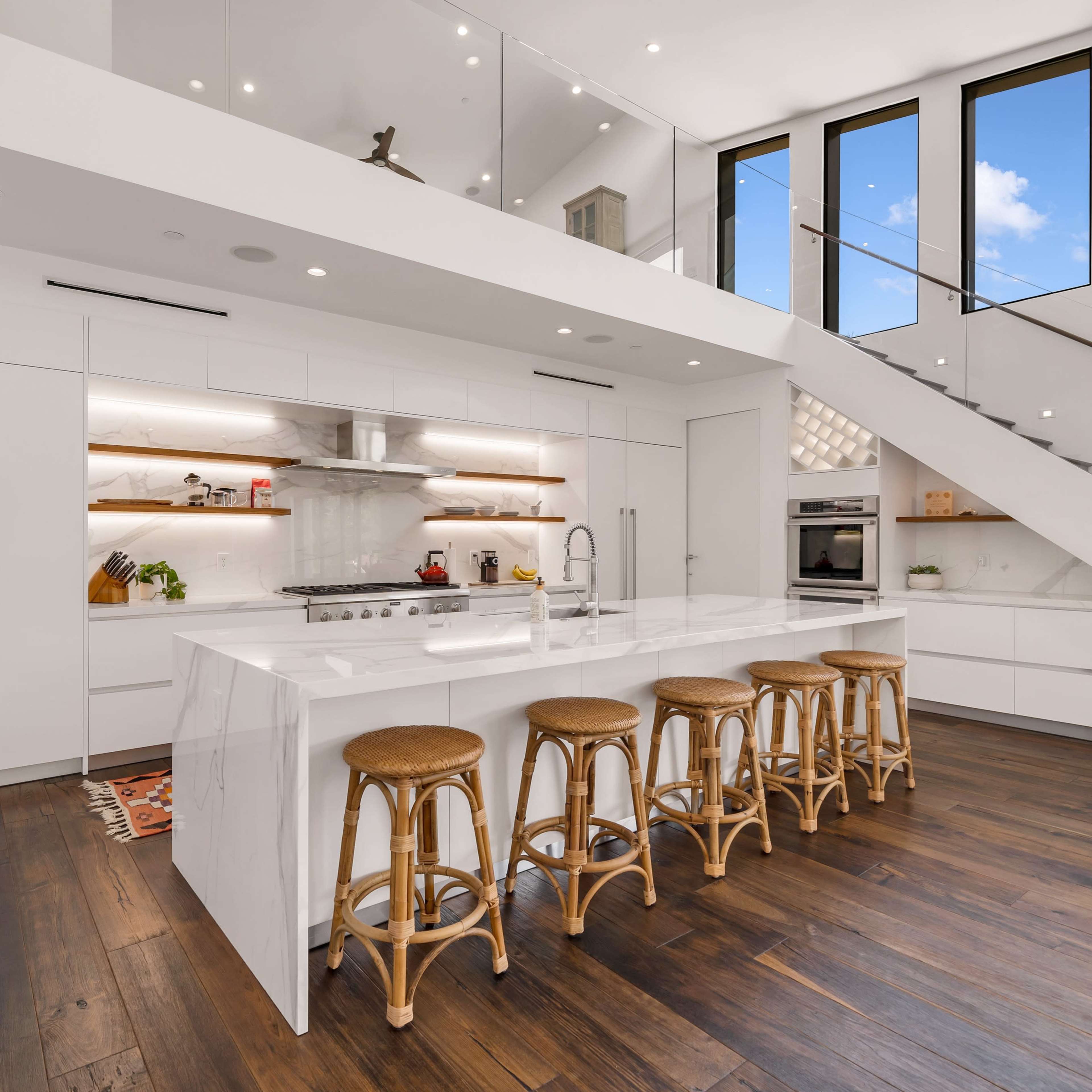 The kitchen features white cabinetry, a large marble island with seating, and wooden floors under ample natural light streaming through tall windows.