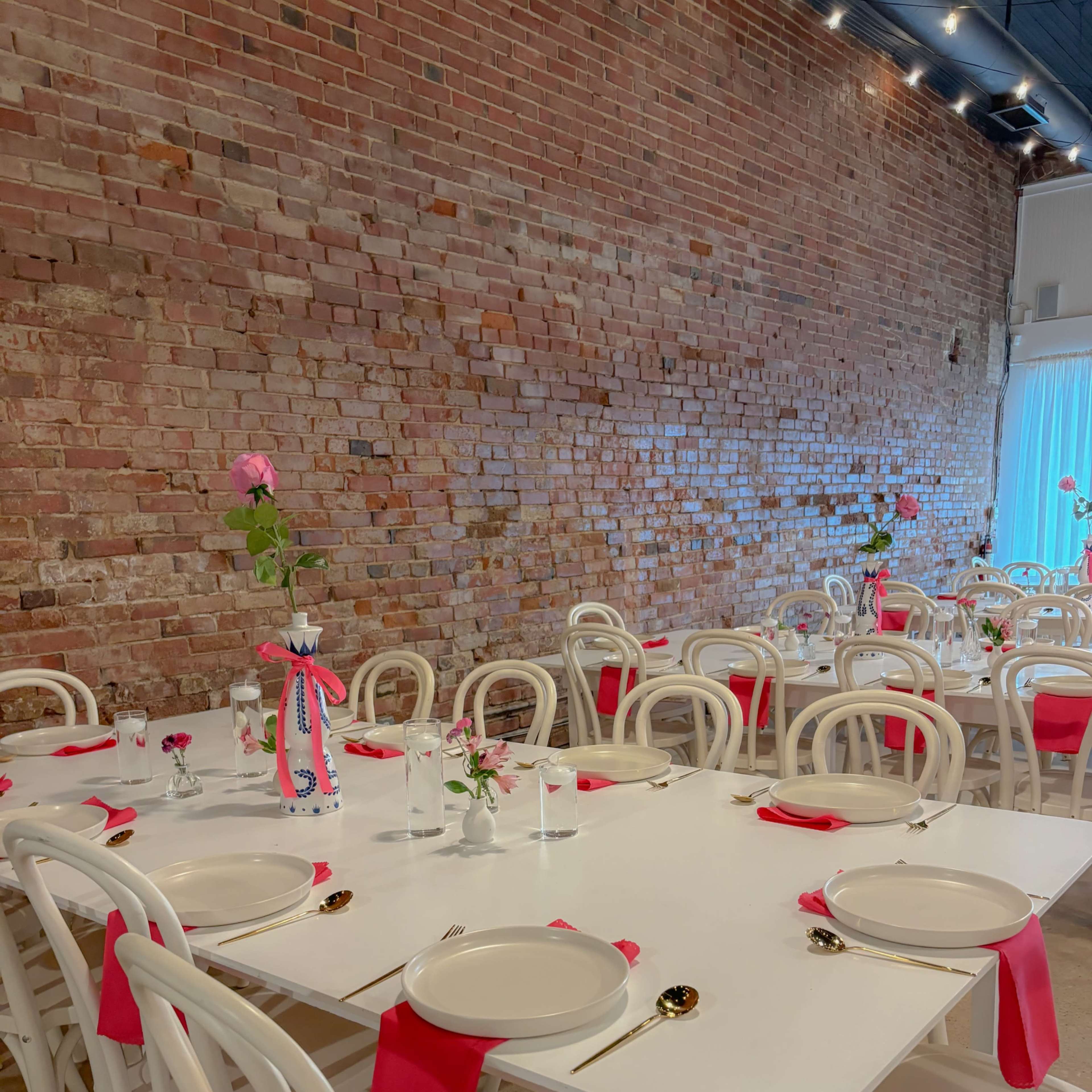 A dining area features several white tables set with plates and red napkins, surrounded by white chairs against a brick wall.