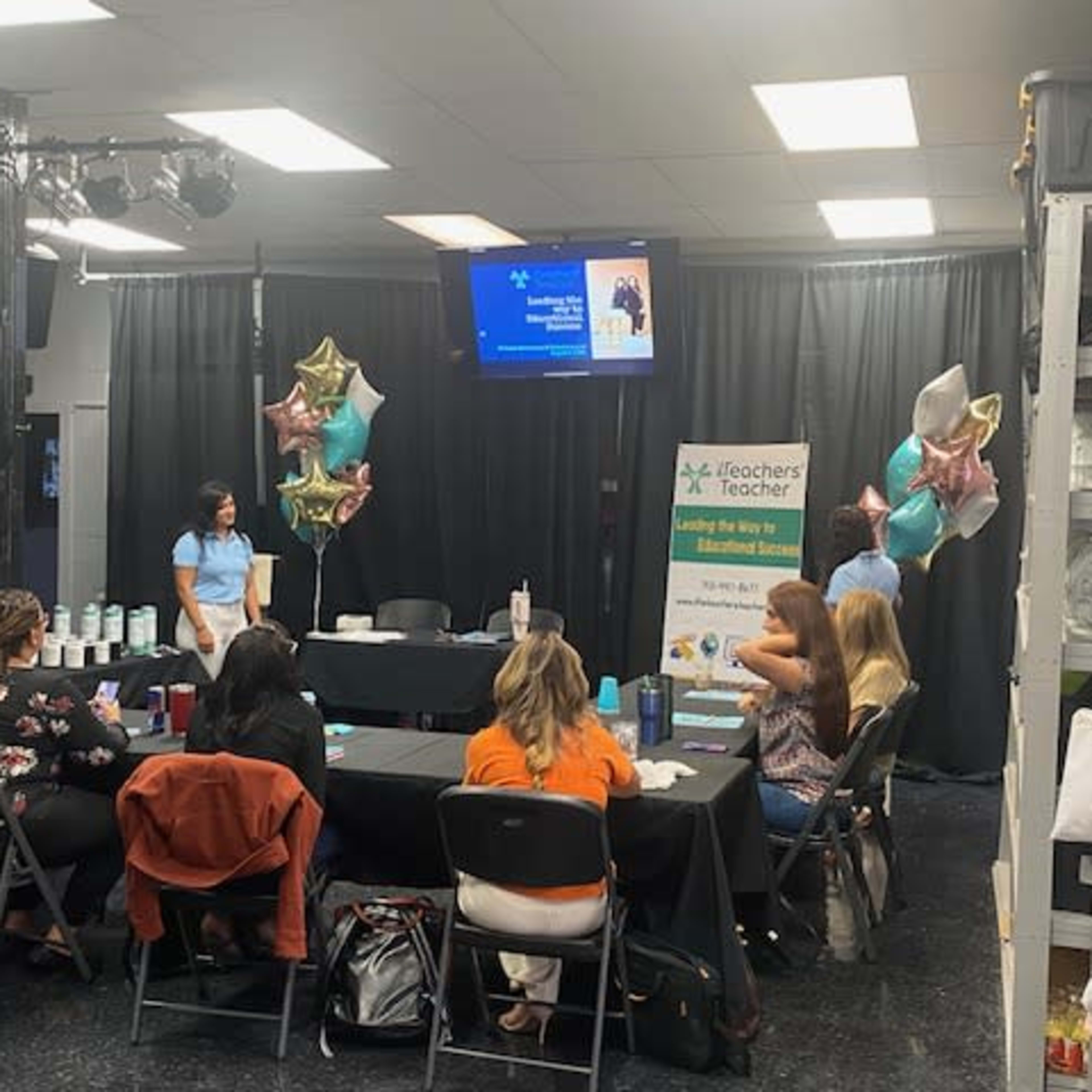A group of people sits at a long black table during a presentation, while balloons and a banner promoting iTeachers are displayed in the background.
