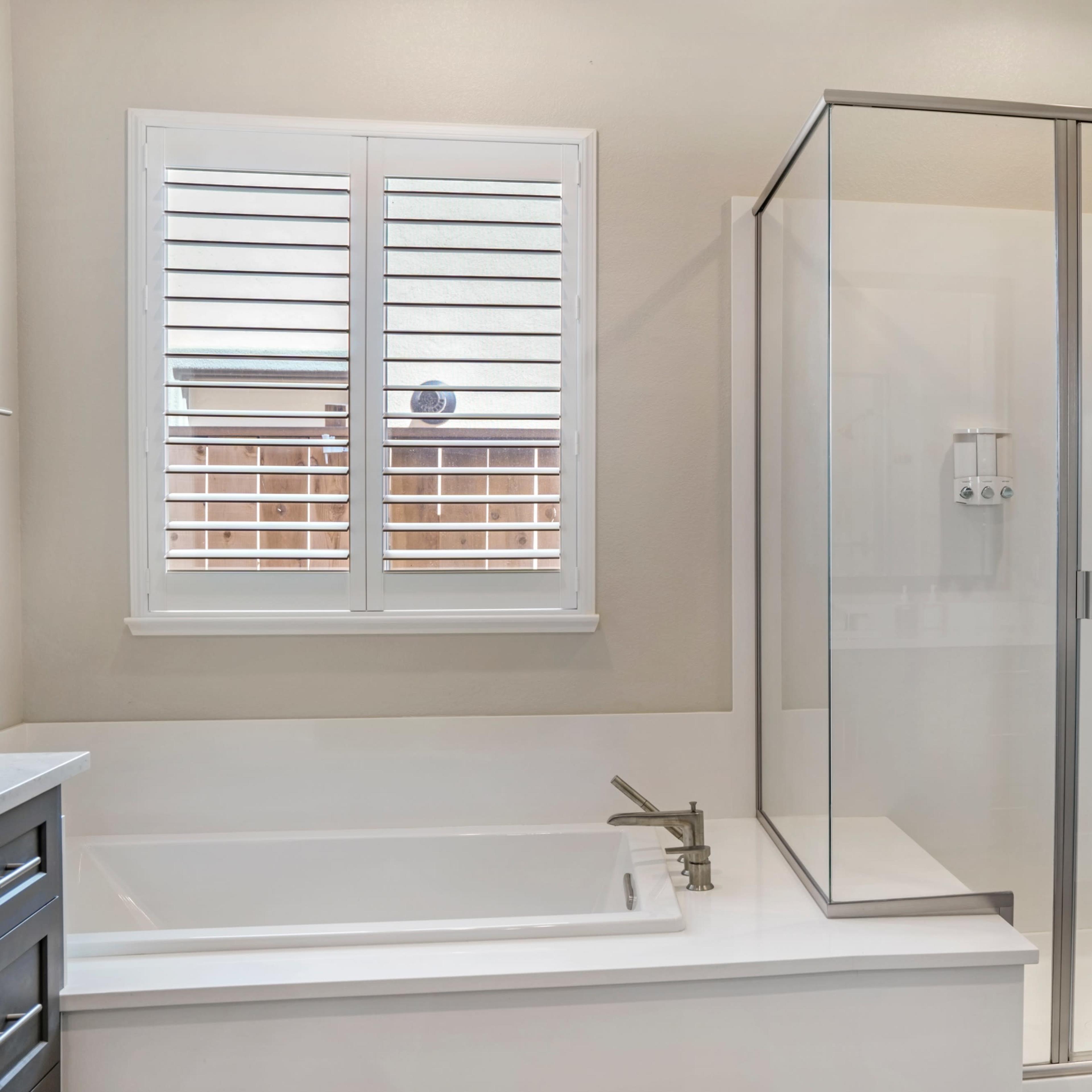A modern bathroom featuring a glass-enclosed shower next to a white bathtub, with a window partially covering a wooden fence outside.