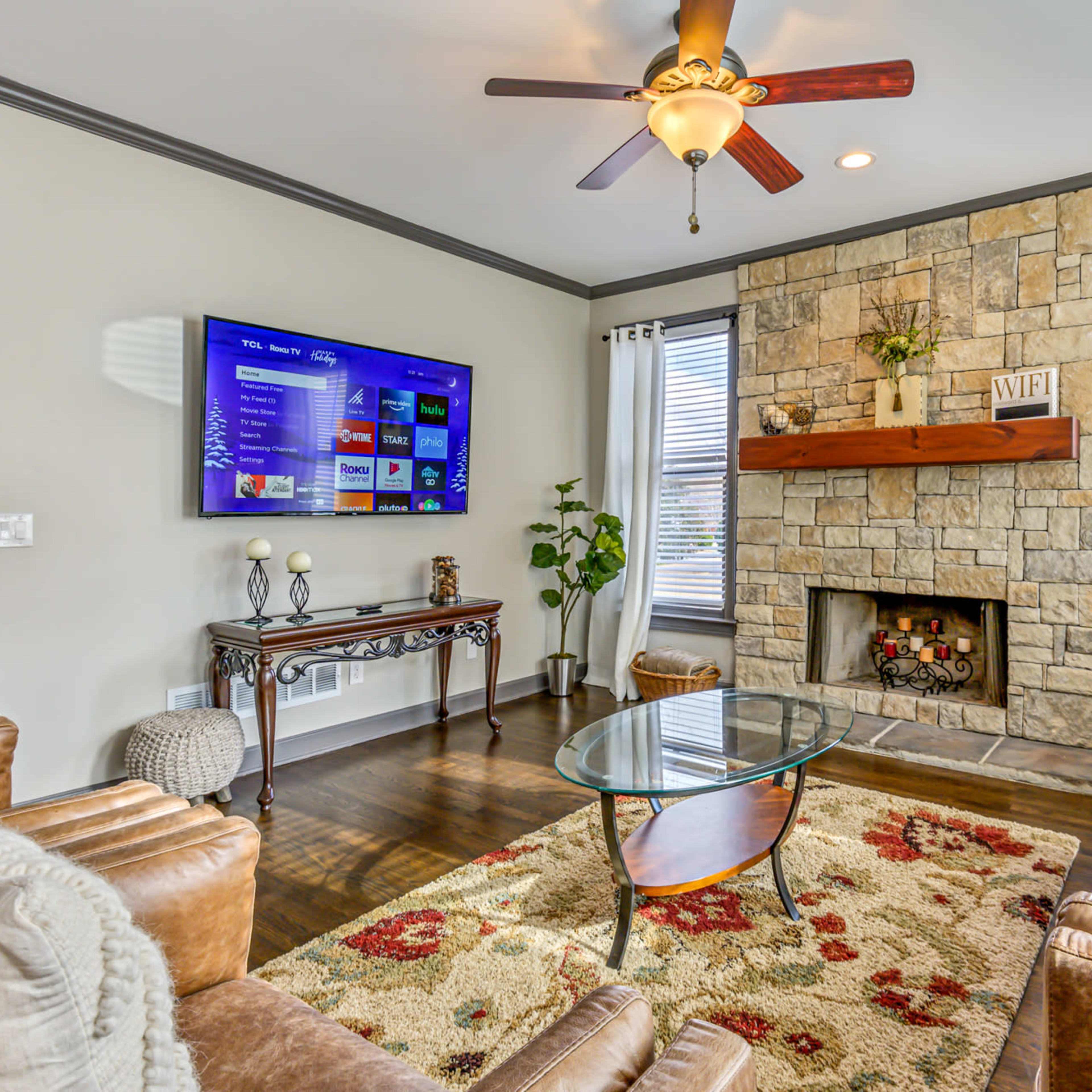 The living room features a stone fireplace, a flat-screen TV mounted on the wall, a glass coffee table, and a sofa set arranged around a patterned area rug.