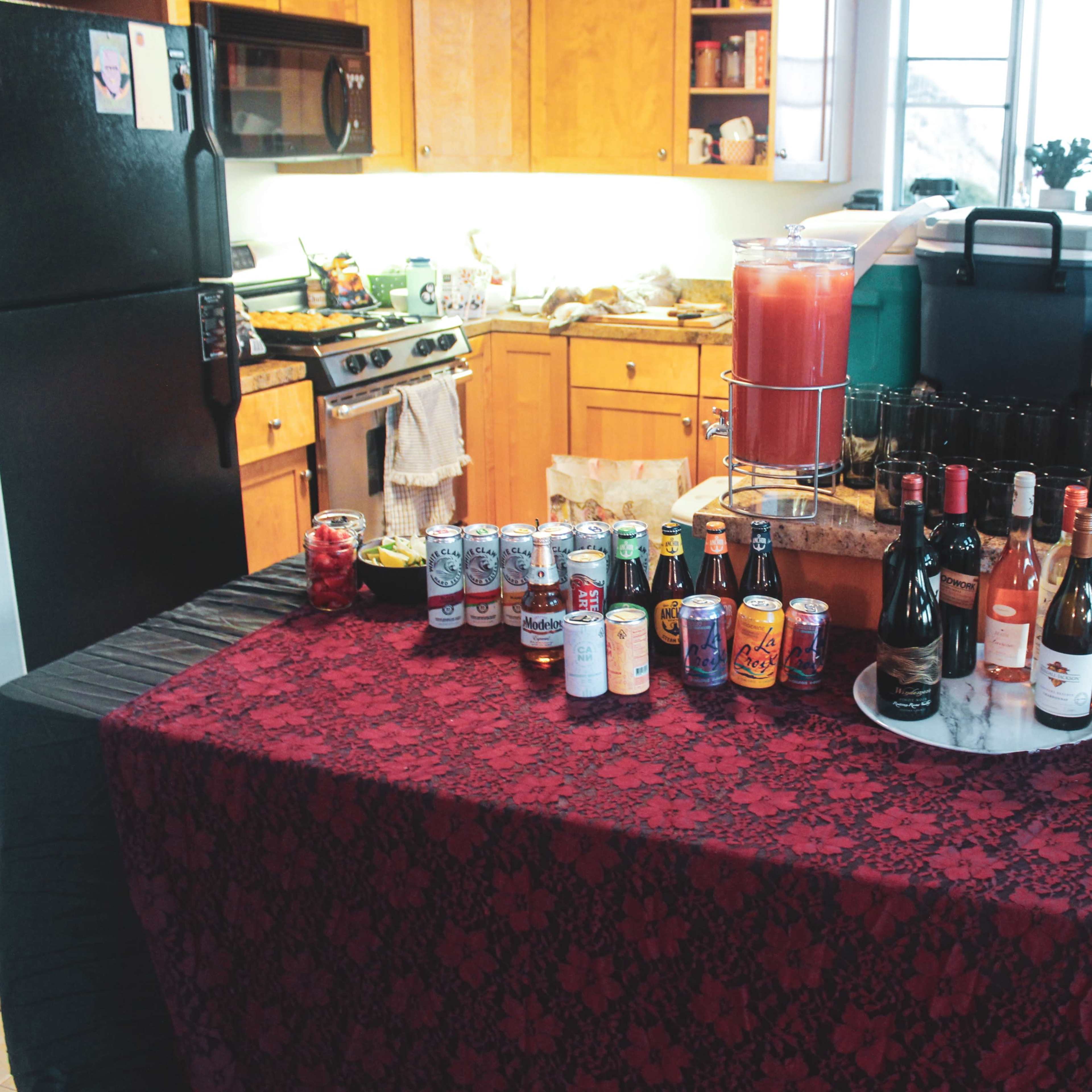 A table covered with a black tablecloth displays an array of drinks, including cans and bottles, in a kitchen setting.