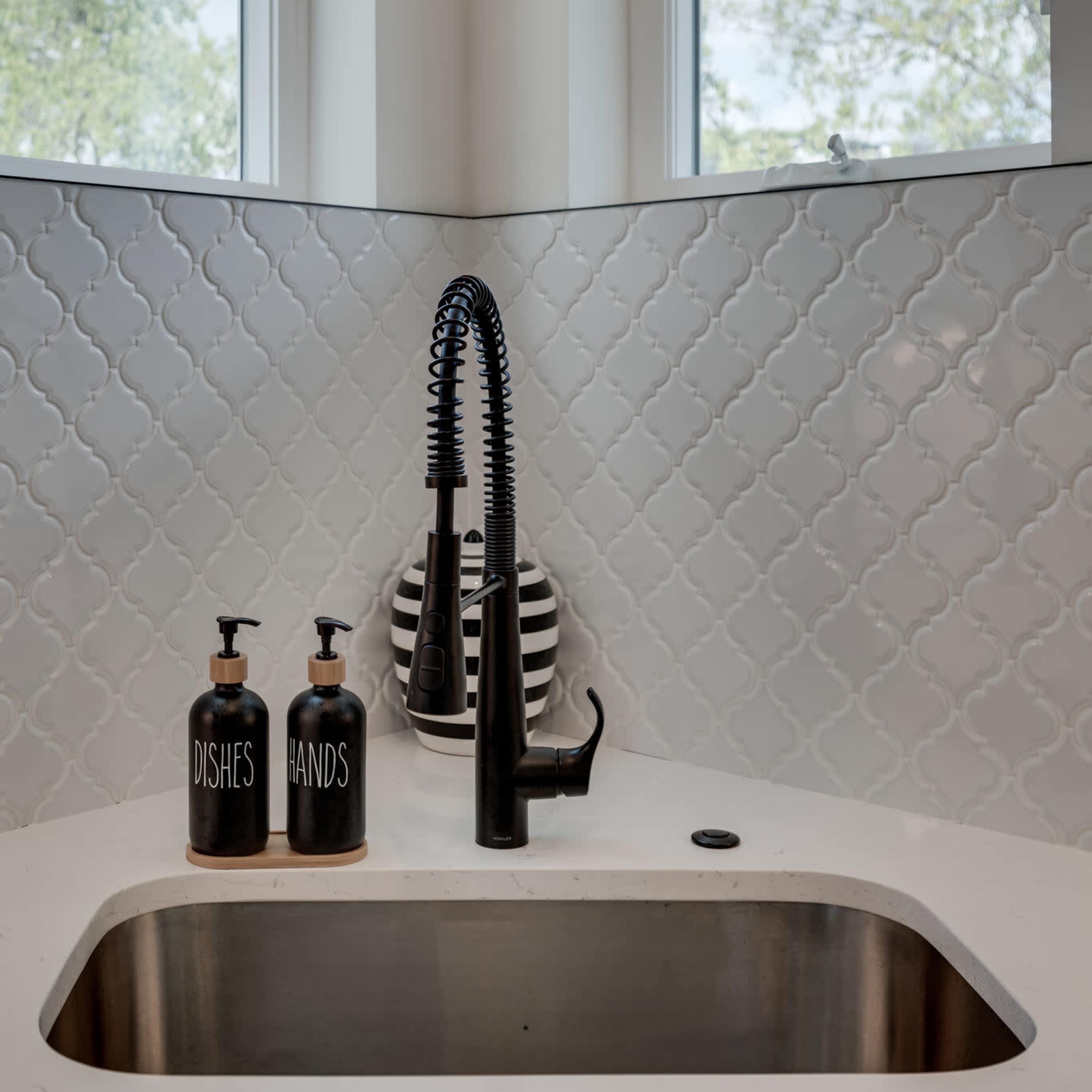 A black faucet is mounted above a stainless steel sink, flanked by two labeled soap dispensers, against a backdrop of patterned white tiles and large windows.
