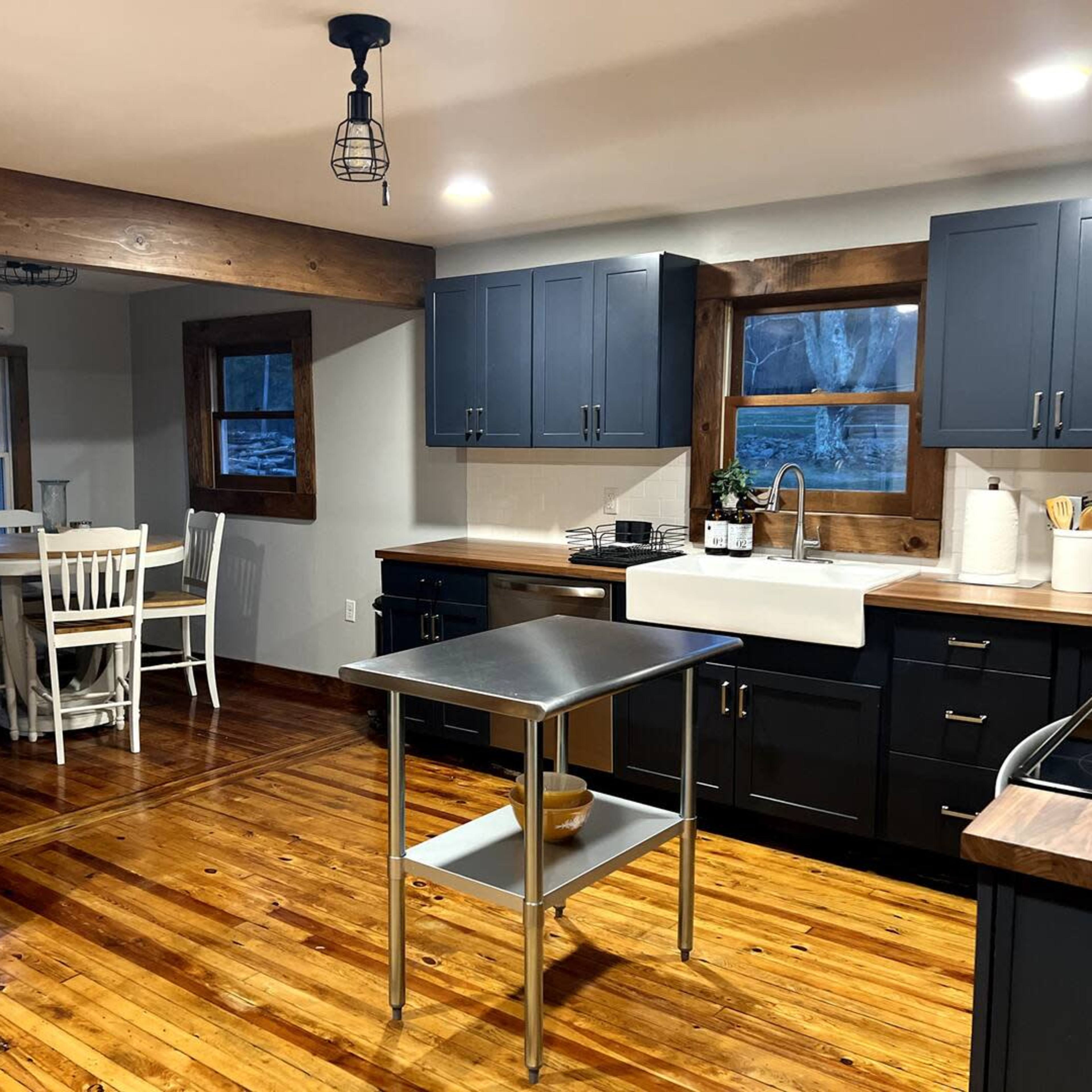 A modern kitchen features dark blue cabinets, a central island with a stainless steel top, and warm wooden floors, with a dining area visible through a window.