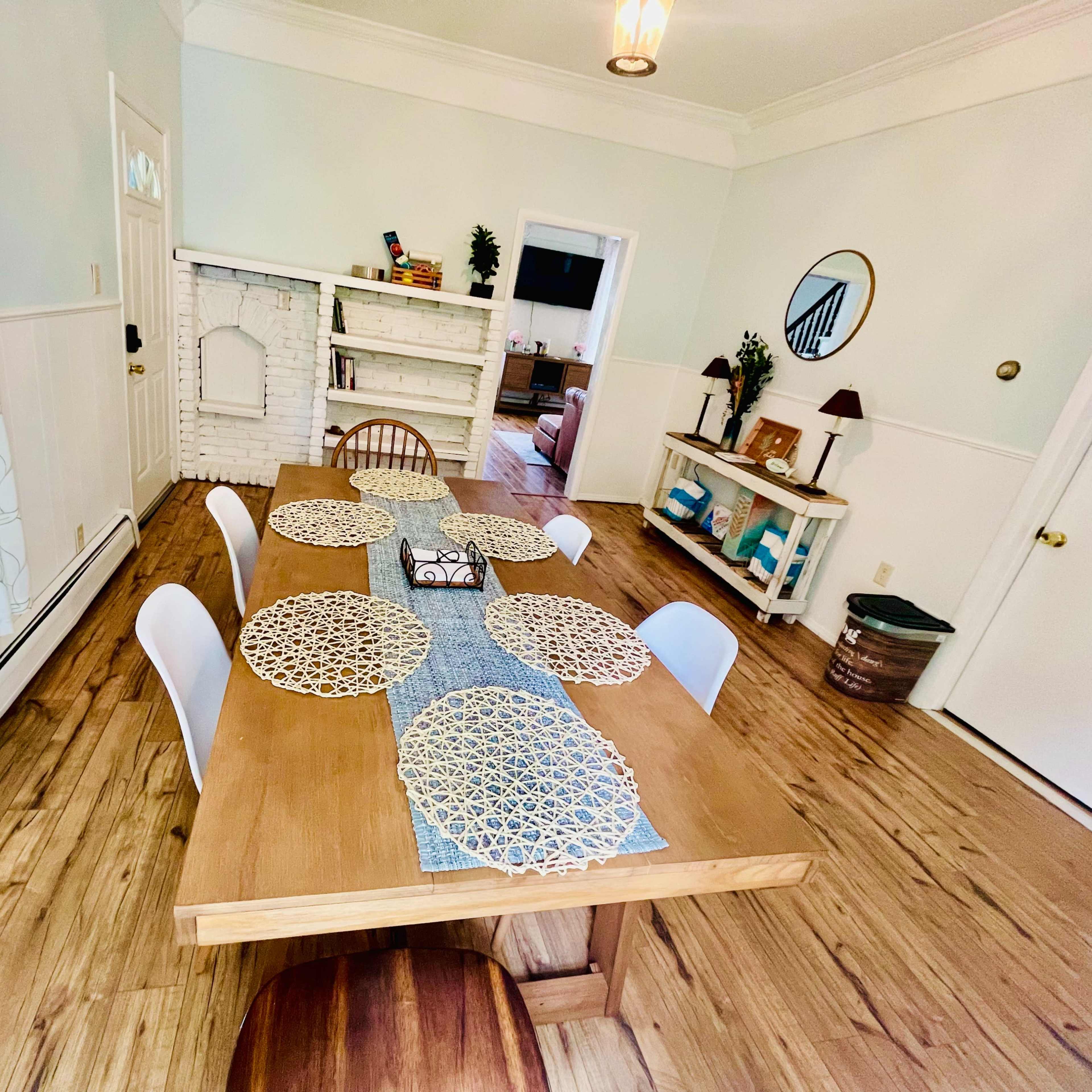 A wooden dining table is set with placemats, and a runner is placed centrally in a light-filled room featuring white walls and hardwood flooring.