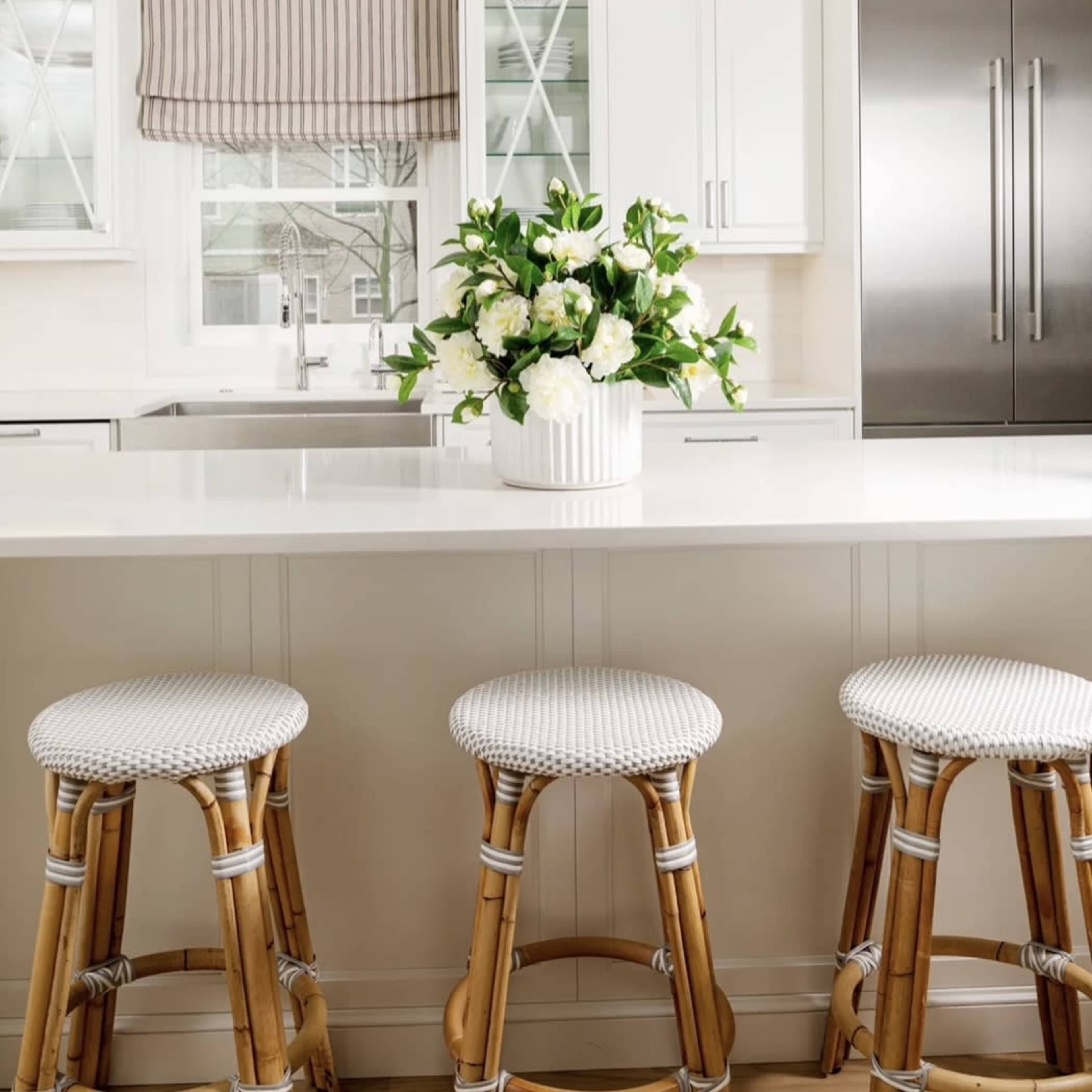 A modern kitchen with white cabinets, a large island topped with a floral centerpiece, and three woven bar stools.