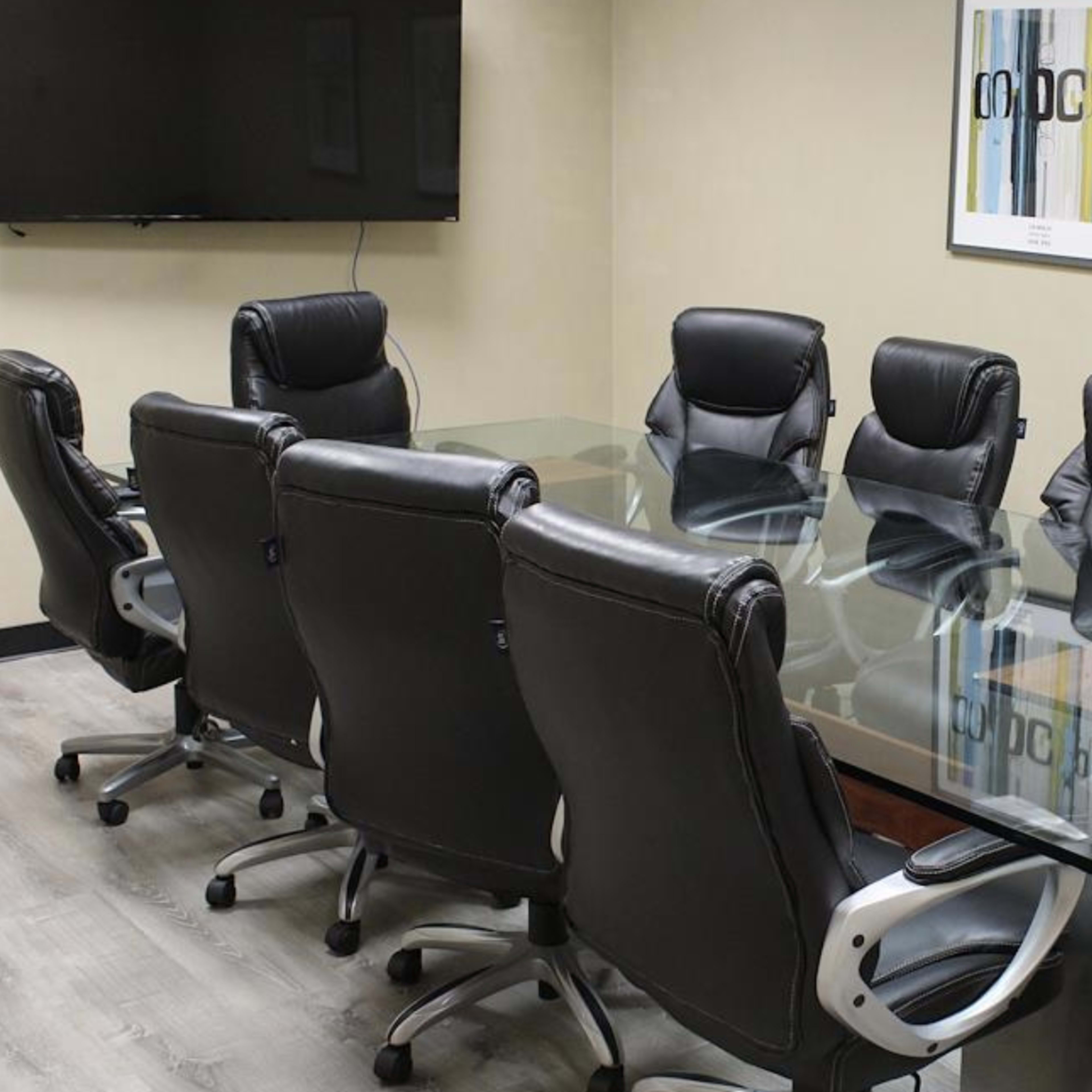 A conference room features a glass table surrounded by eight black ergonomic chairs, with a television mounted on the wall and framed art displayed.