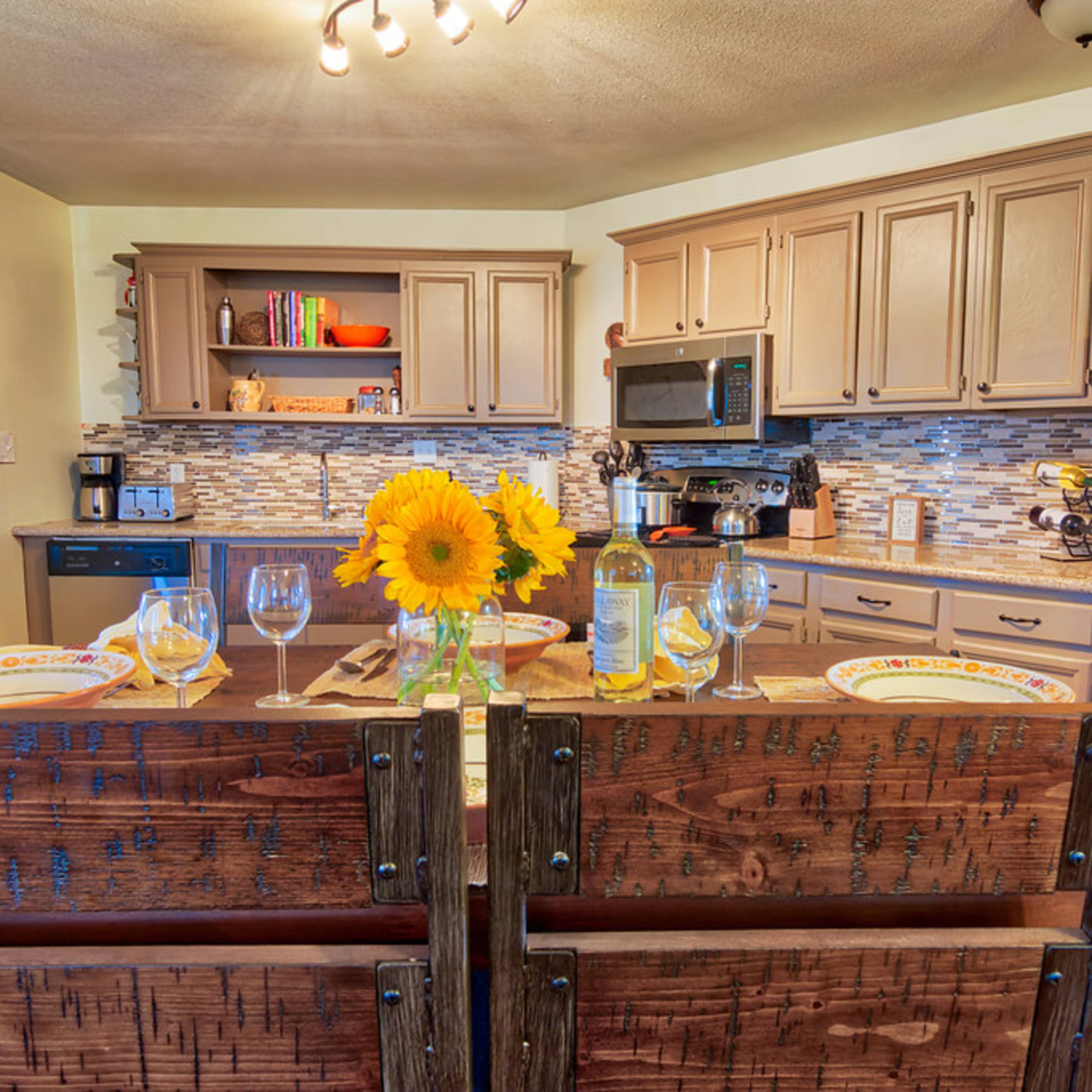A kitchen features a wooden dining table set with plates and glasses, adorned with a vase of sunflowers, while cabinets and appliances are neatly arranged along the walls.