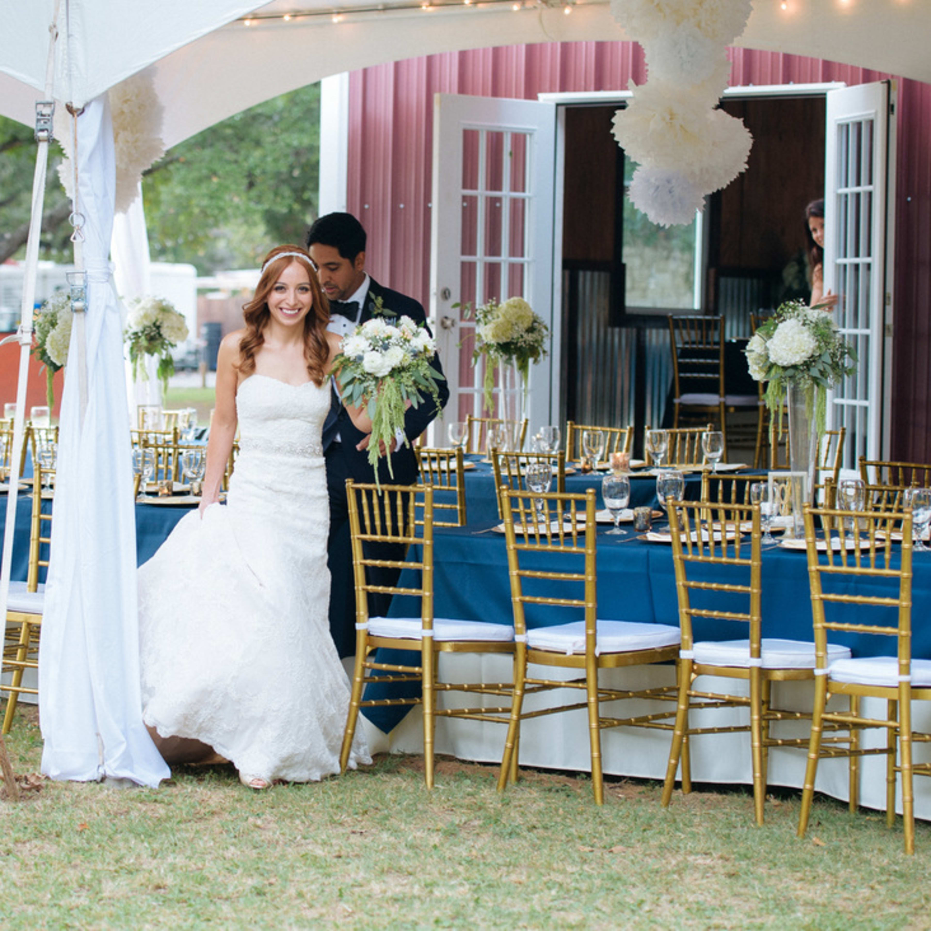 A bride in a white dress walks into a tented reception area with a groom, surrounded by elegantly arranged tables and golden chairs.