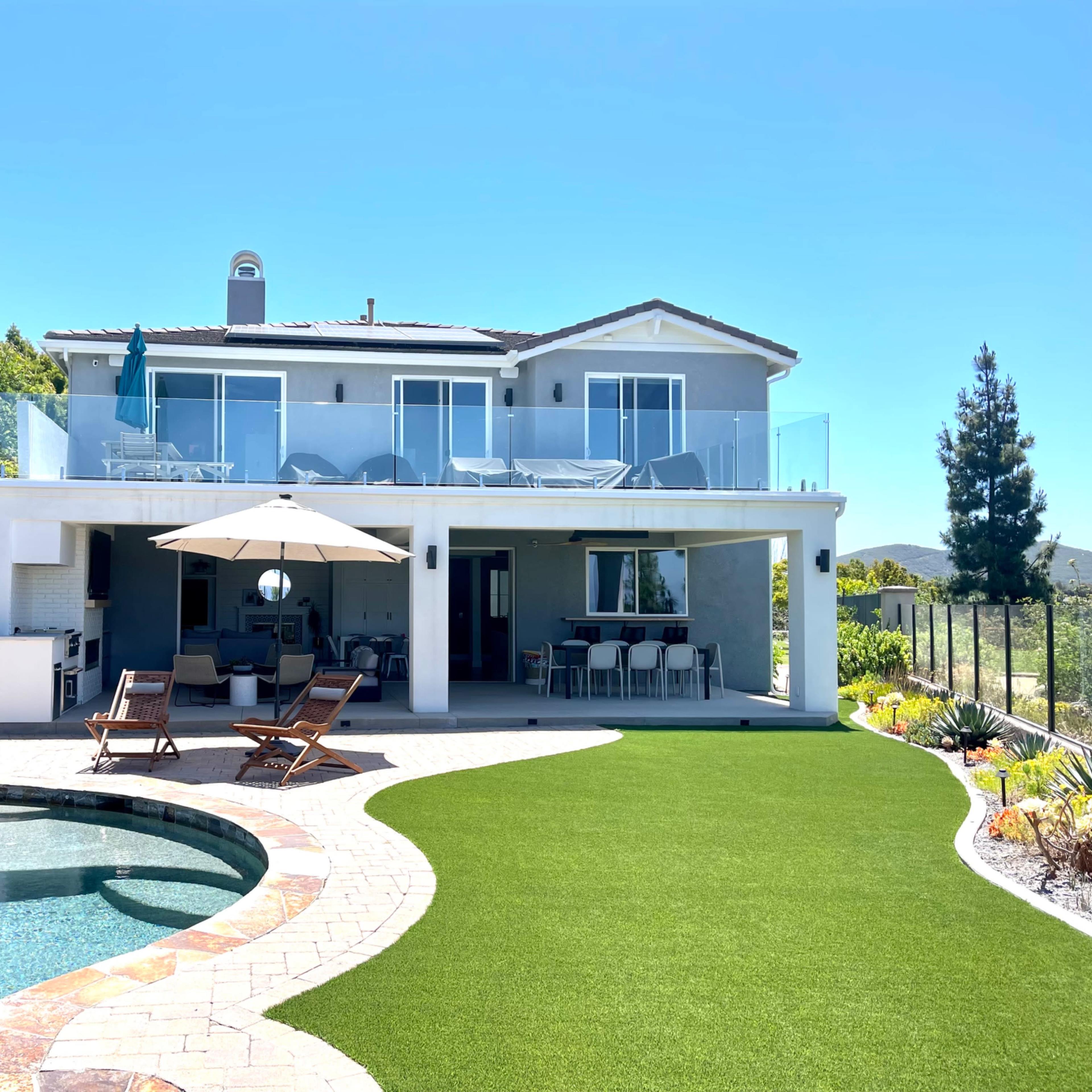 A two-story house with a patio overlooking a pool, surrounded by landscaped greenery.
