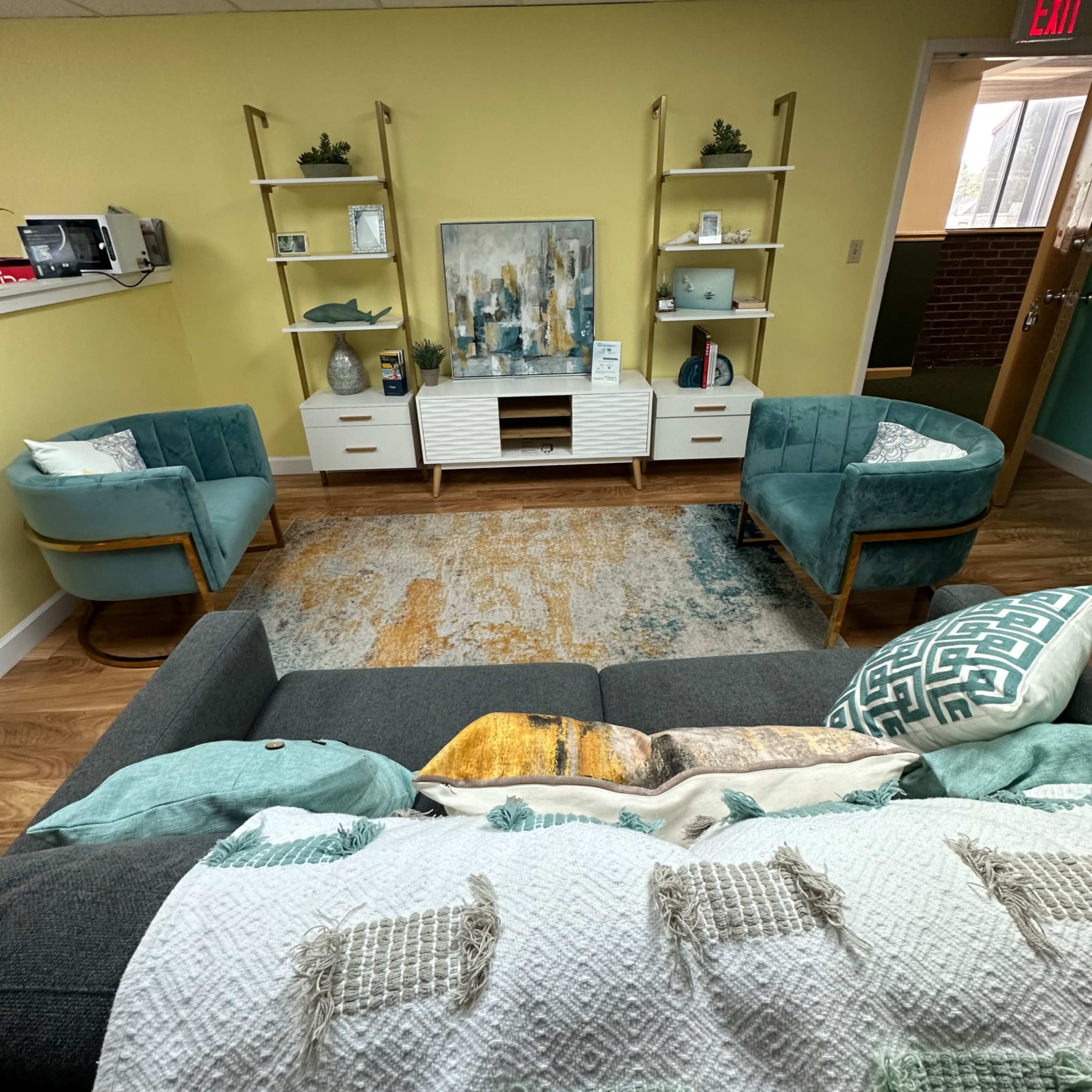 A modern living area featuring a gray sofa, two teal armchairs, a rug, a media console with shelves, and a decorative wall painting.