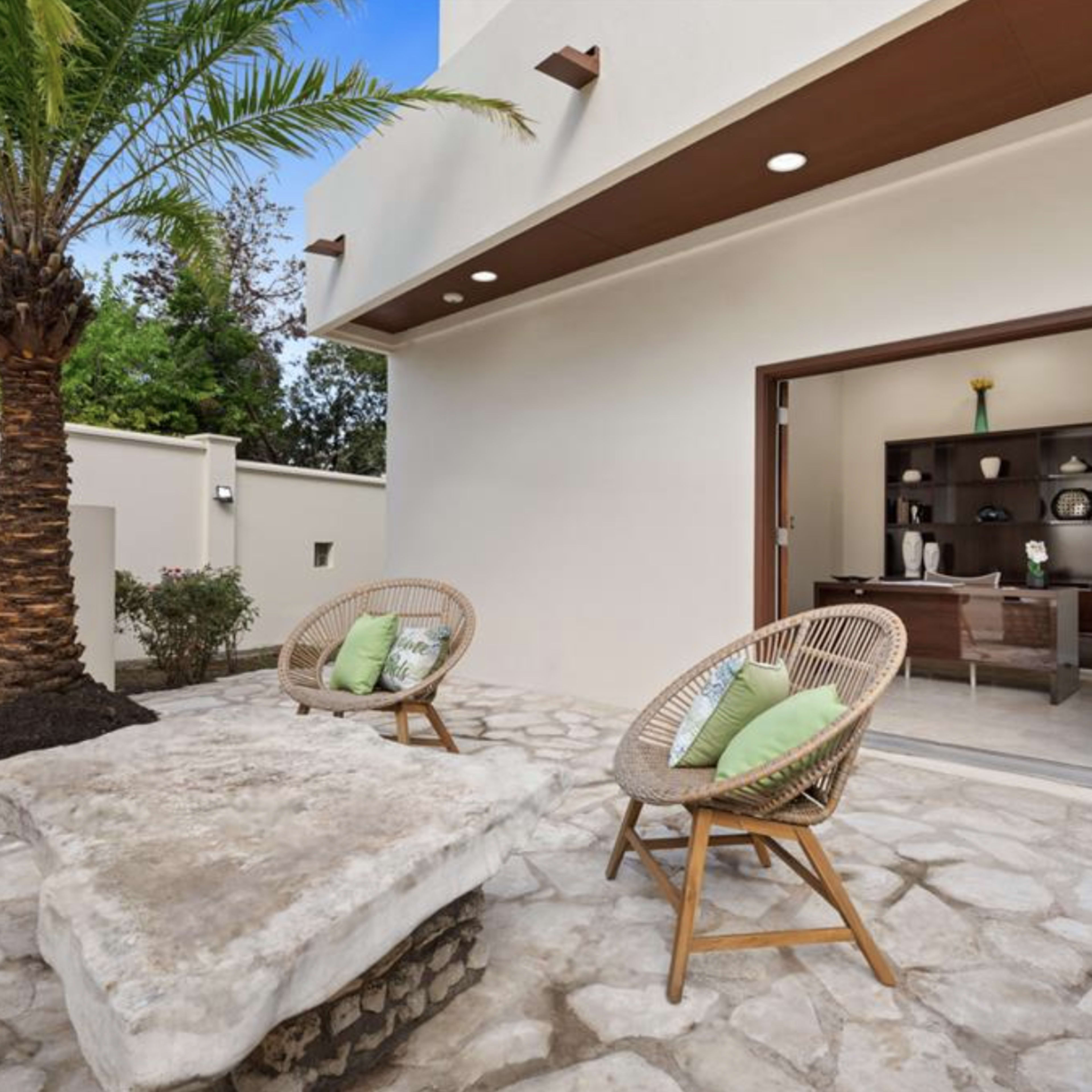 A modern outdoor seating area with two rattan chairs and a stone table beside a palm tree.