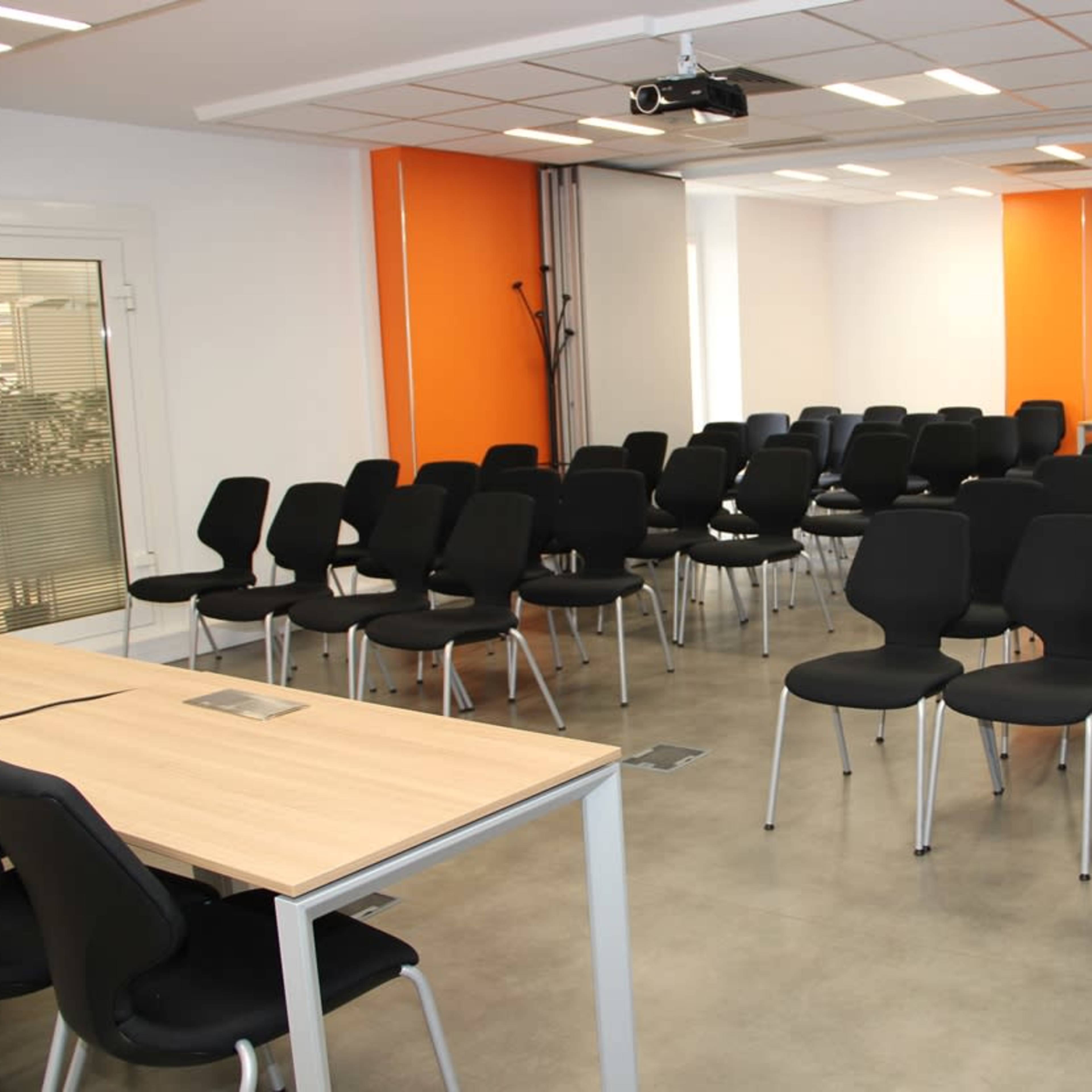A conference room set up with rows of black chairs facing a desk and projector.
