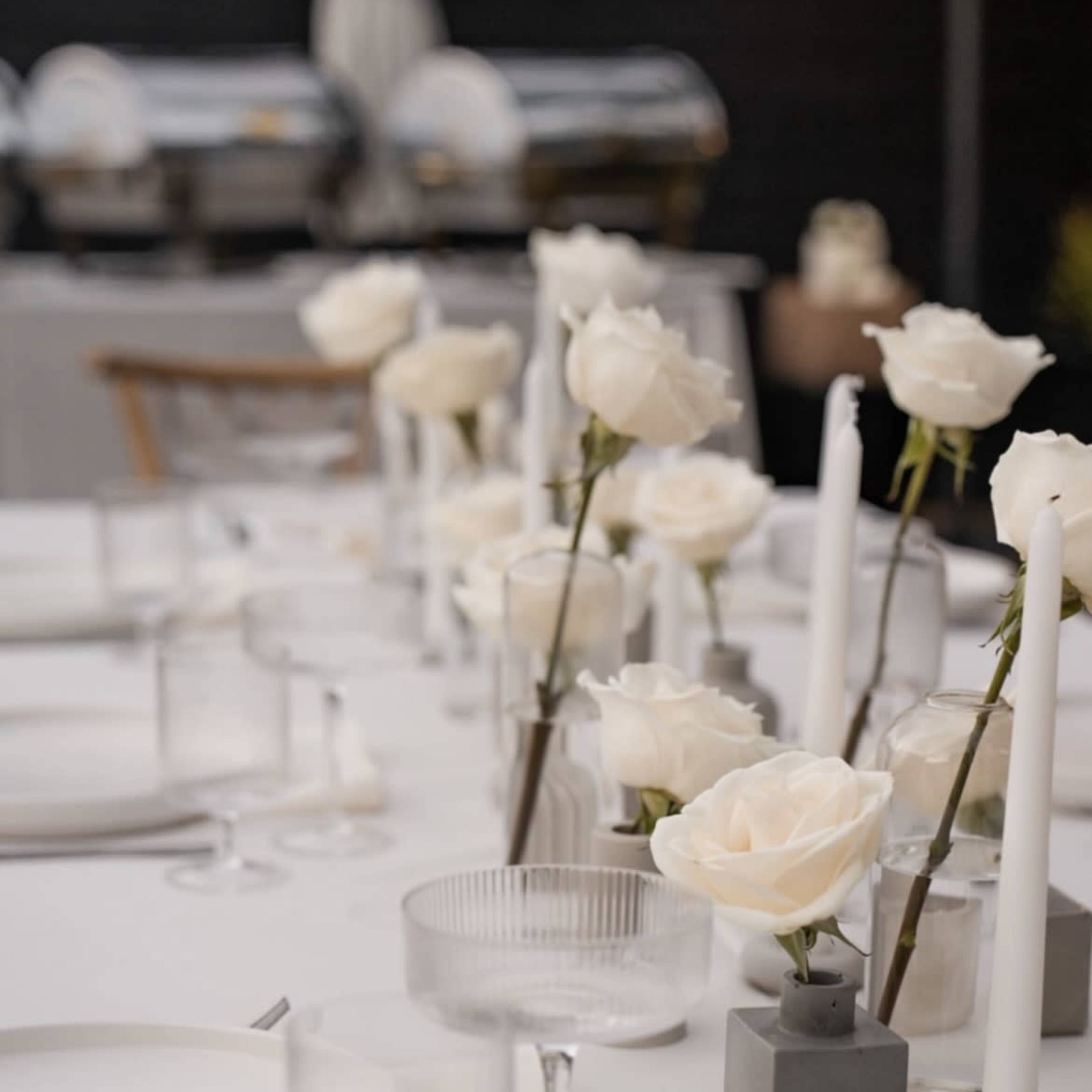 A long dining table is set with white dishes, clear glasses, and centerpieces of white roses alongside white candles in minimalist holders.